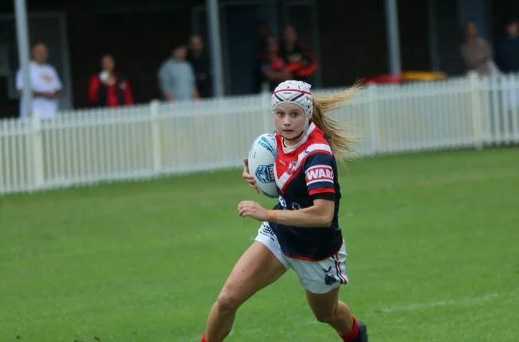 A young girl playing rugby outdoors on a grassy field, holding a white rugby ball, with a white picket fence and spectators in the background.
