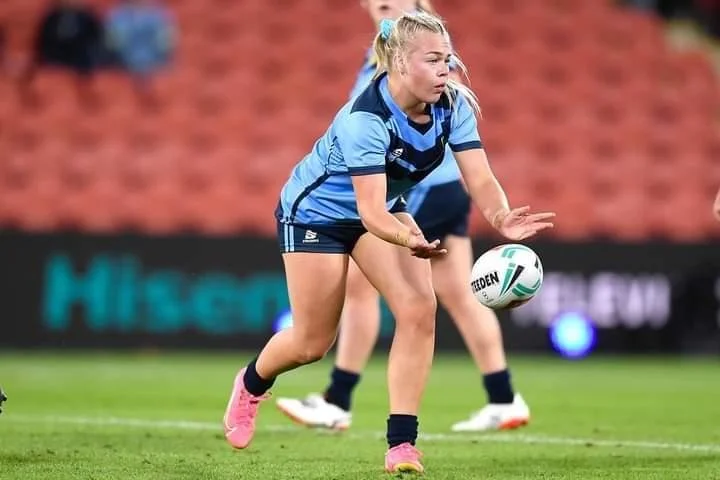 Rugby player in blue jersey catching a rugby ball on field, with stadium seating in background.
