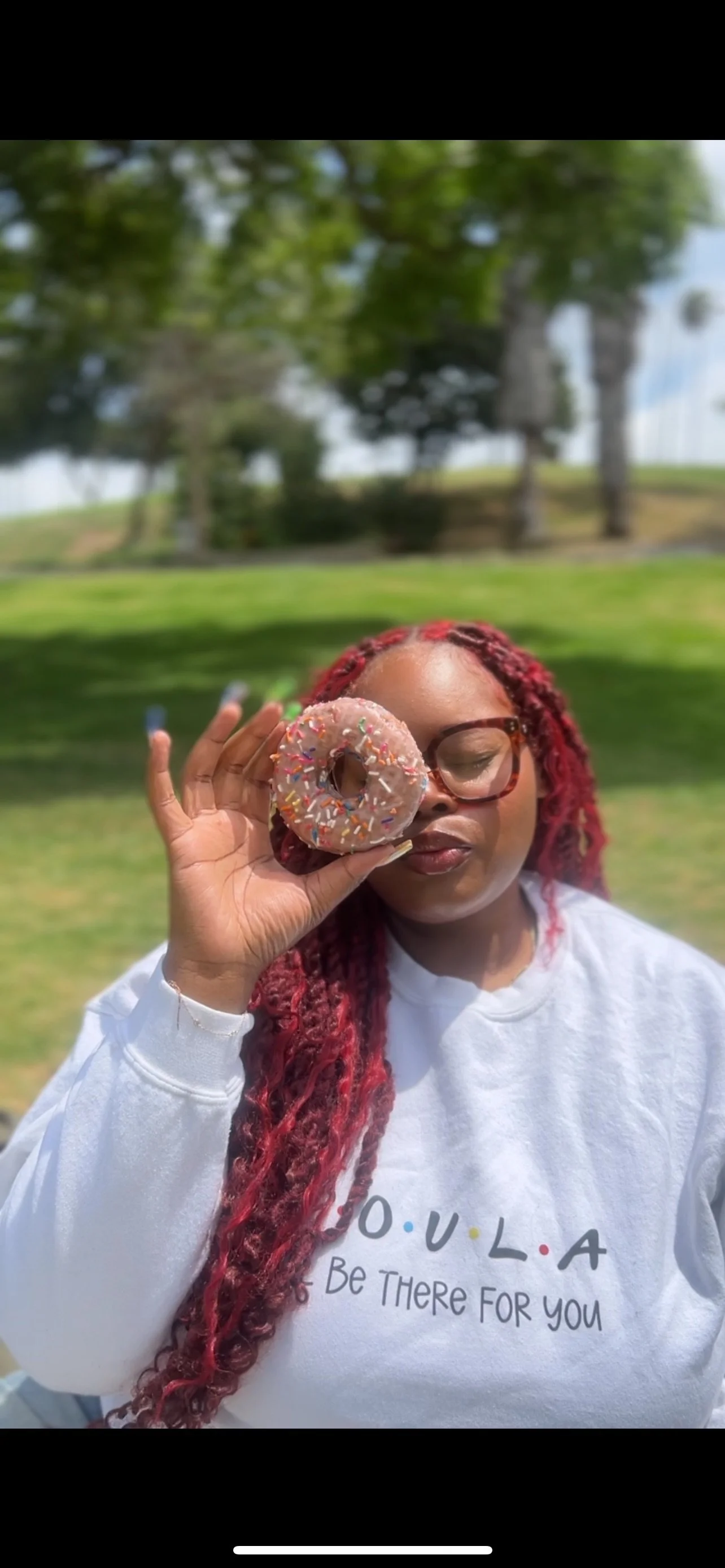 A woman with red braids and glasses holds a pink sprinkled donut in front of her eye outdoors, with trees and grass in the background.