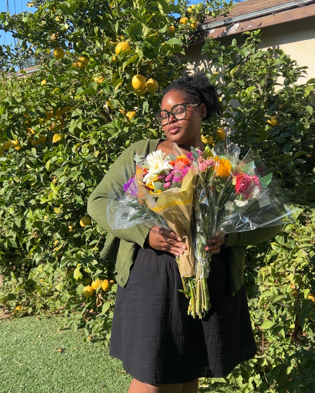 A woman with glasses holding a large bouquet of colorful flowers standing in a garden with lemon trees.