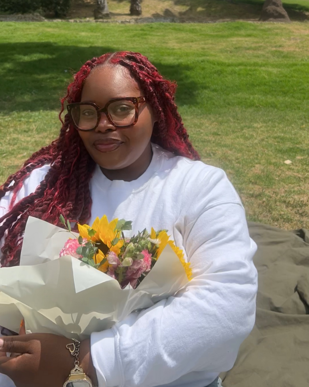 A woman with red curly hair and glasses holding a bouquet of yellow, pink, and purple flowers outdoors on a grassy lawn.