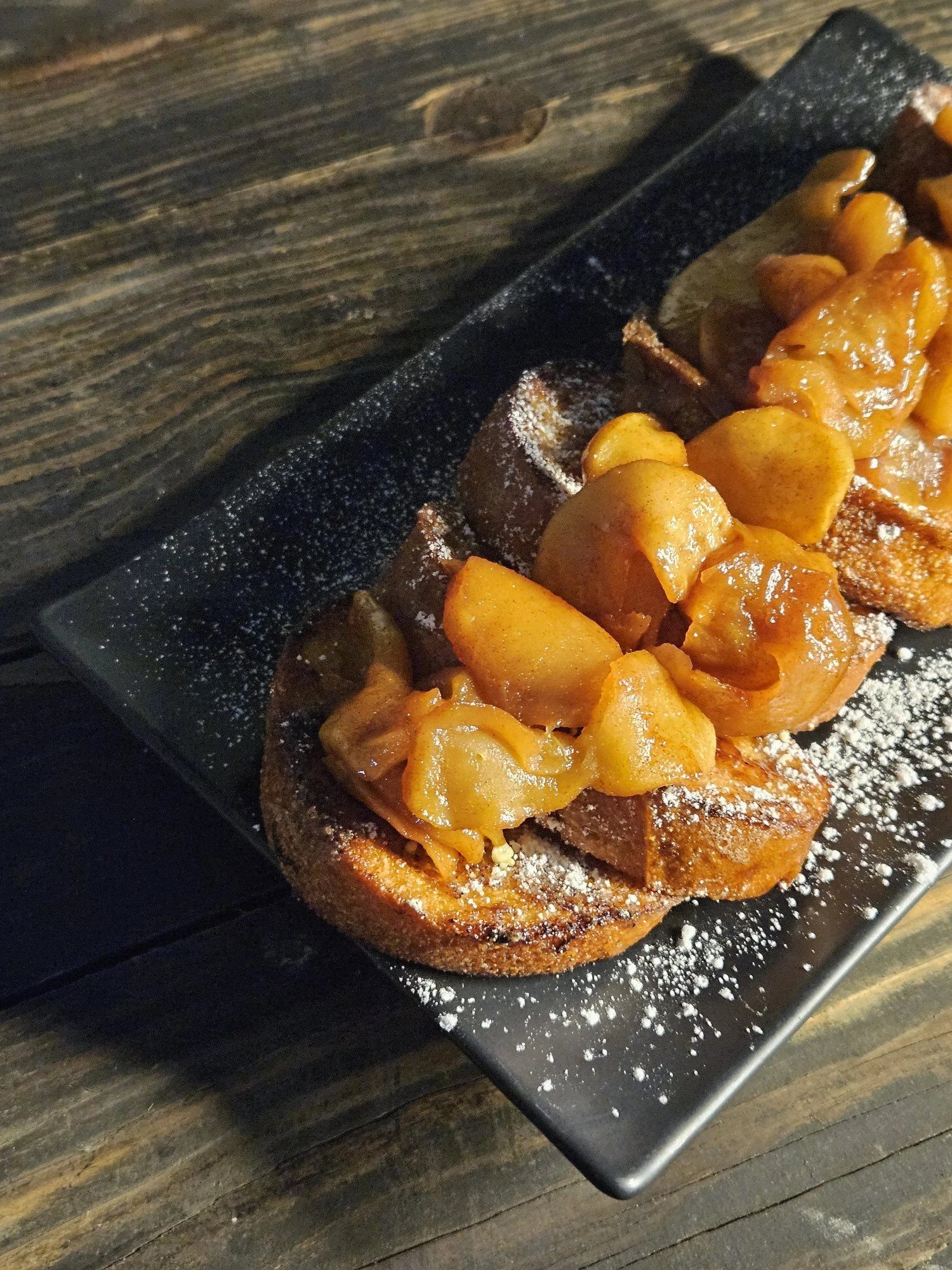 Slices of French toast topped with peach slices, dusted with powdered sugar, on a black rectangular plate.