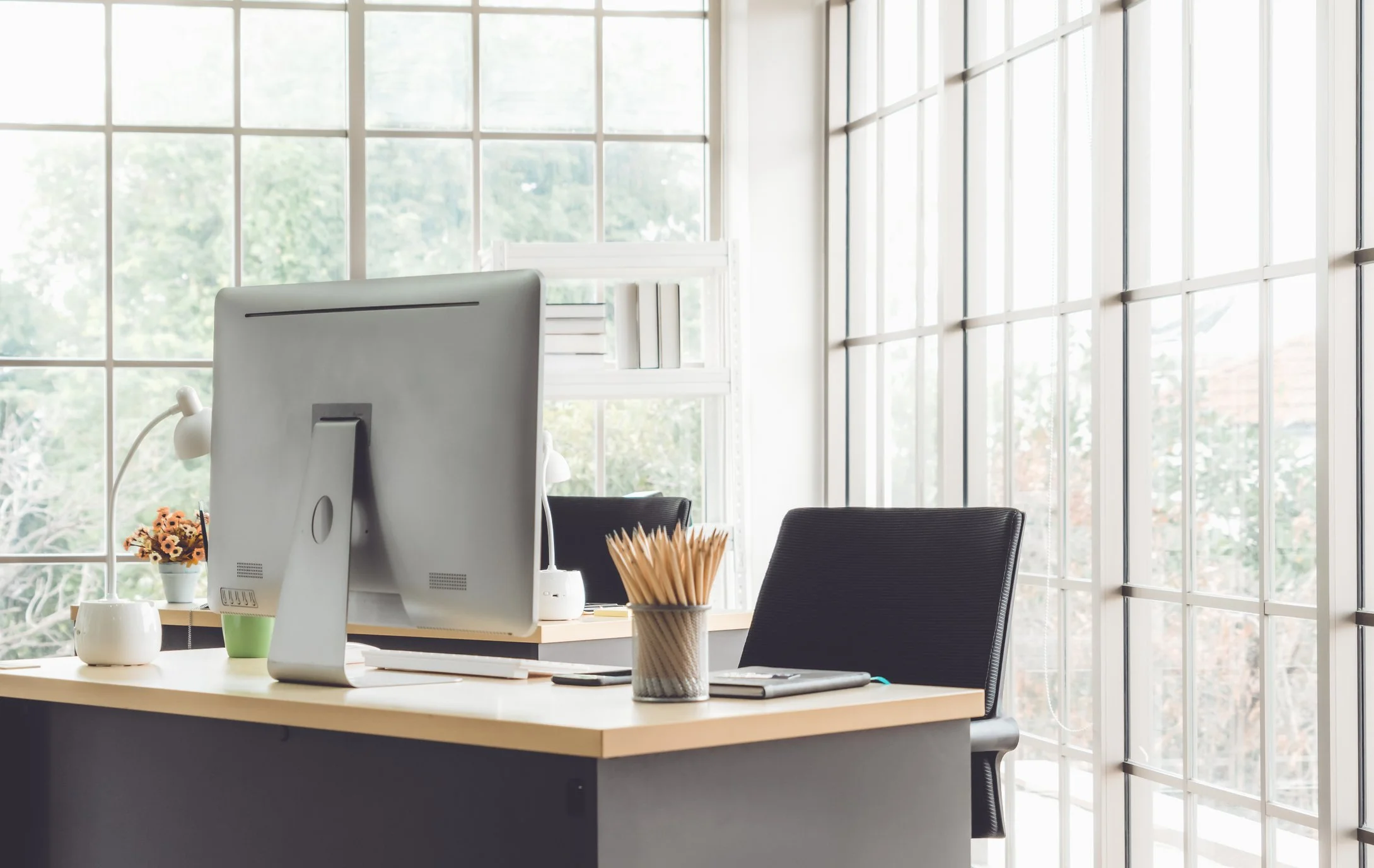 Office workspace with a desk, a large computer monitor, a black chair, a cup filled with pencils, and a windowed wall allowing natural light.