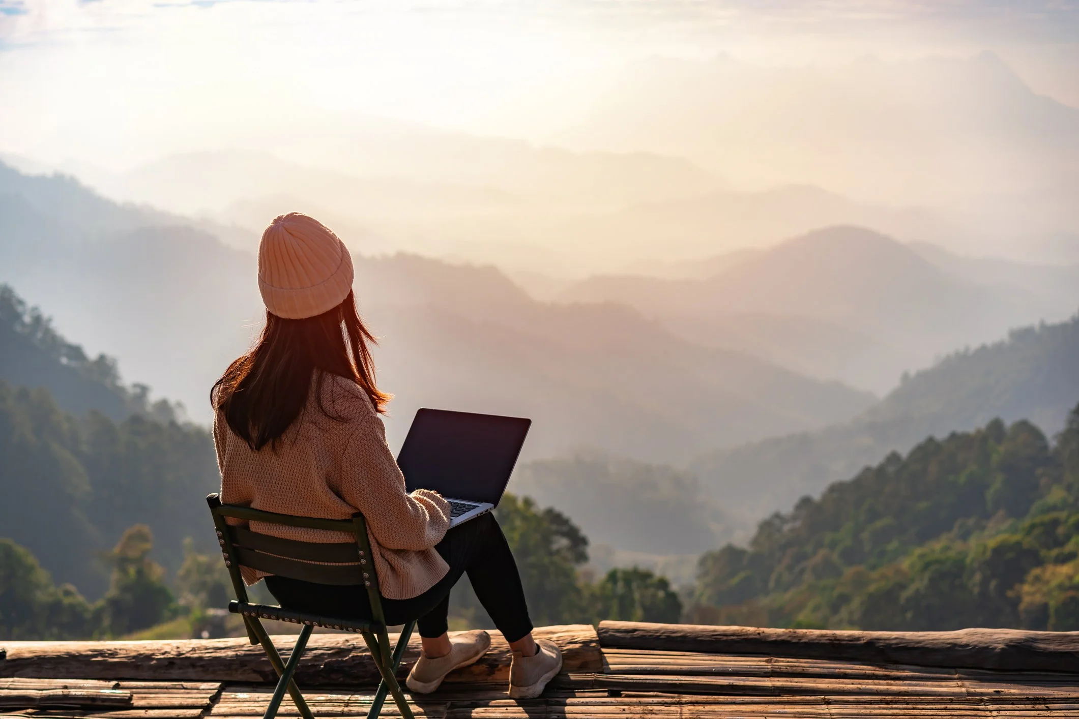 A woman sitting on a wooden deck with a laptop, overlooking a misty mountain landscape at sunrise or sunset.