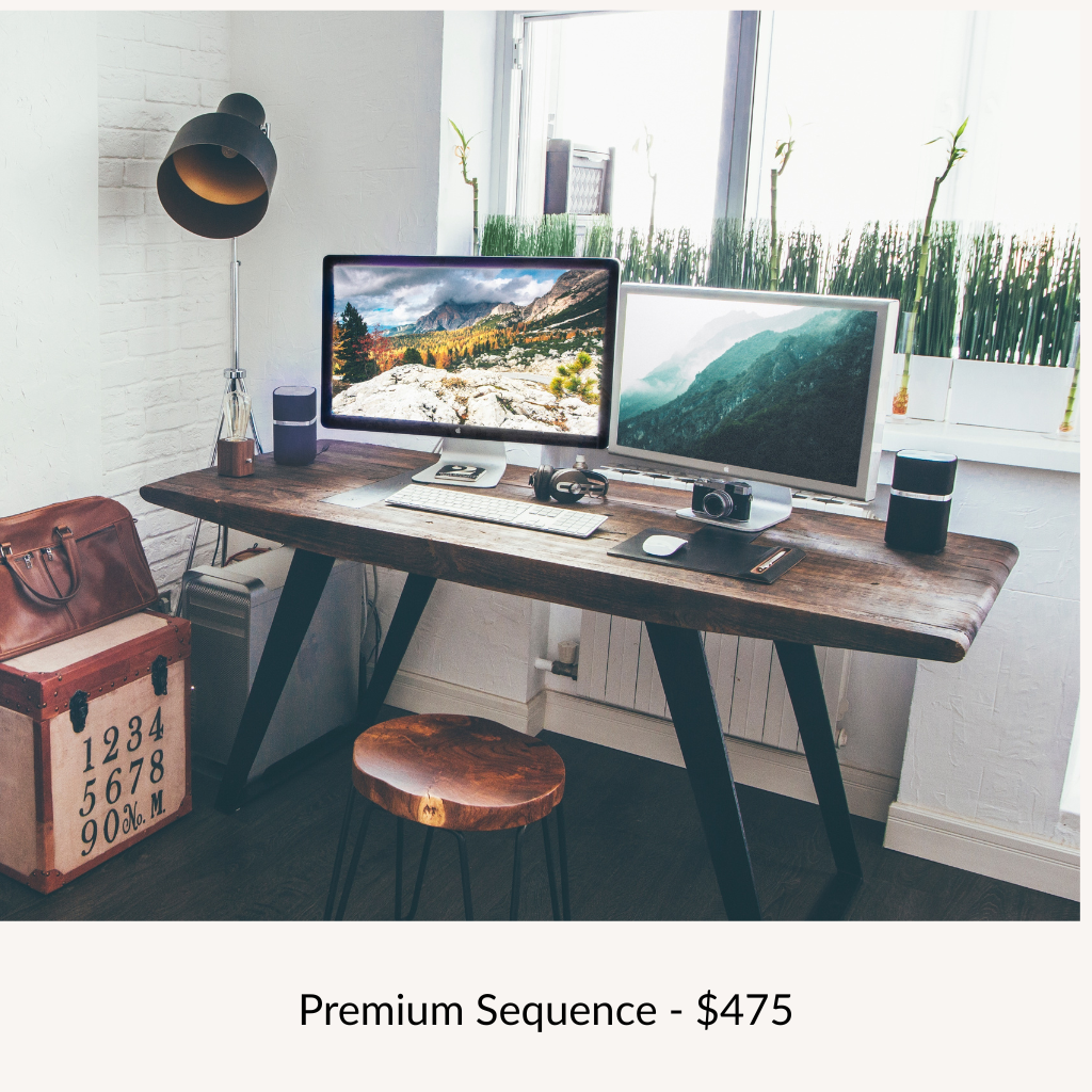 A modern home office desk with two large monitors, a keyboard, mouse, headphones, camera, and a small speaker. There is a wooden stool, a desk lamp, a brown leather bag, and a vintage-style box with numbers and letters. Behind the desk, there is a window with green plants and tall grass outside.