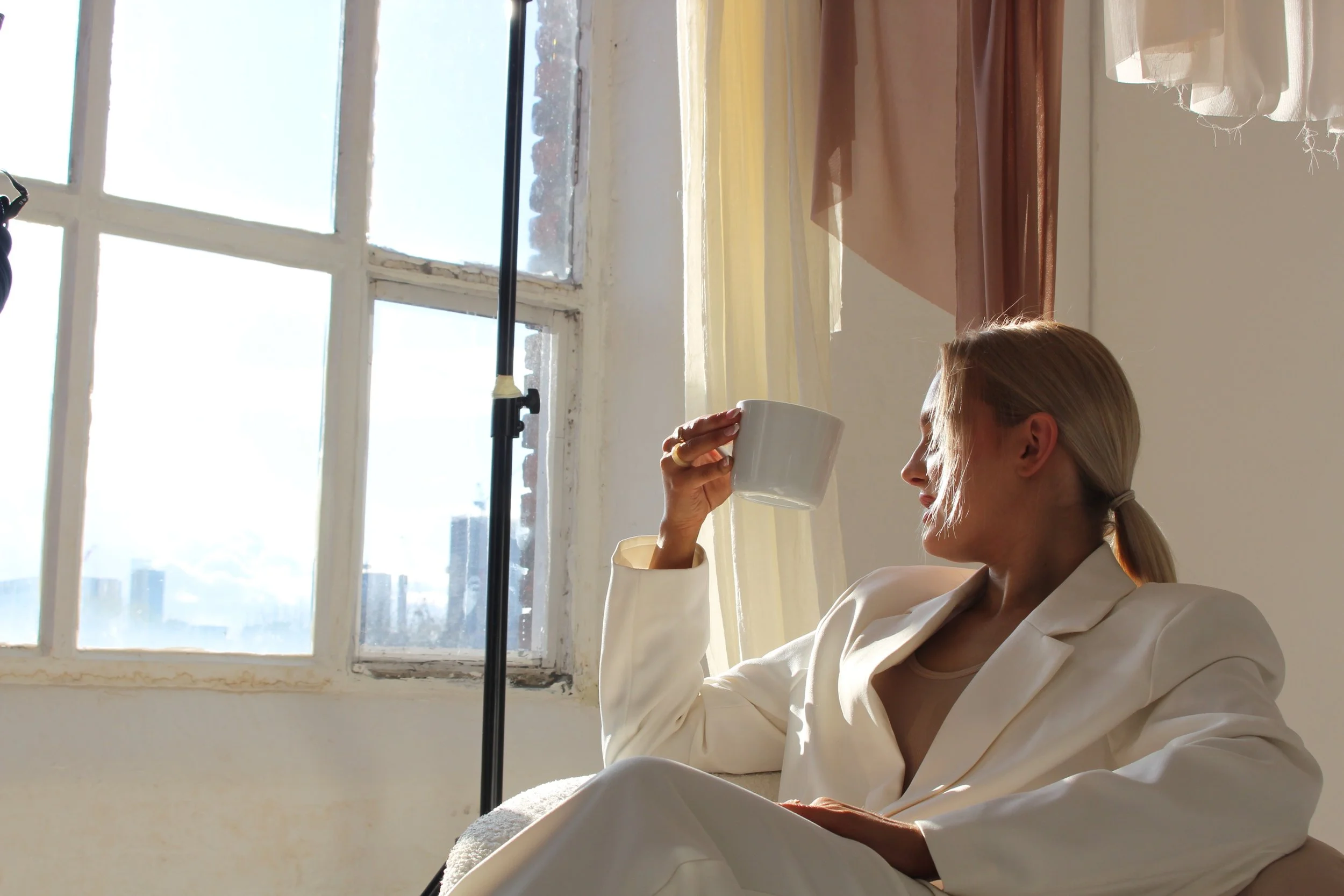 A woman with blonde hair tied back, dressed in a white blazer and beige top, sitting and smiling while drinking from a white mug near a window with sheer curtains. The sun is shining through the window, creating a bright effect.