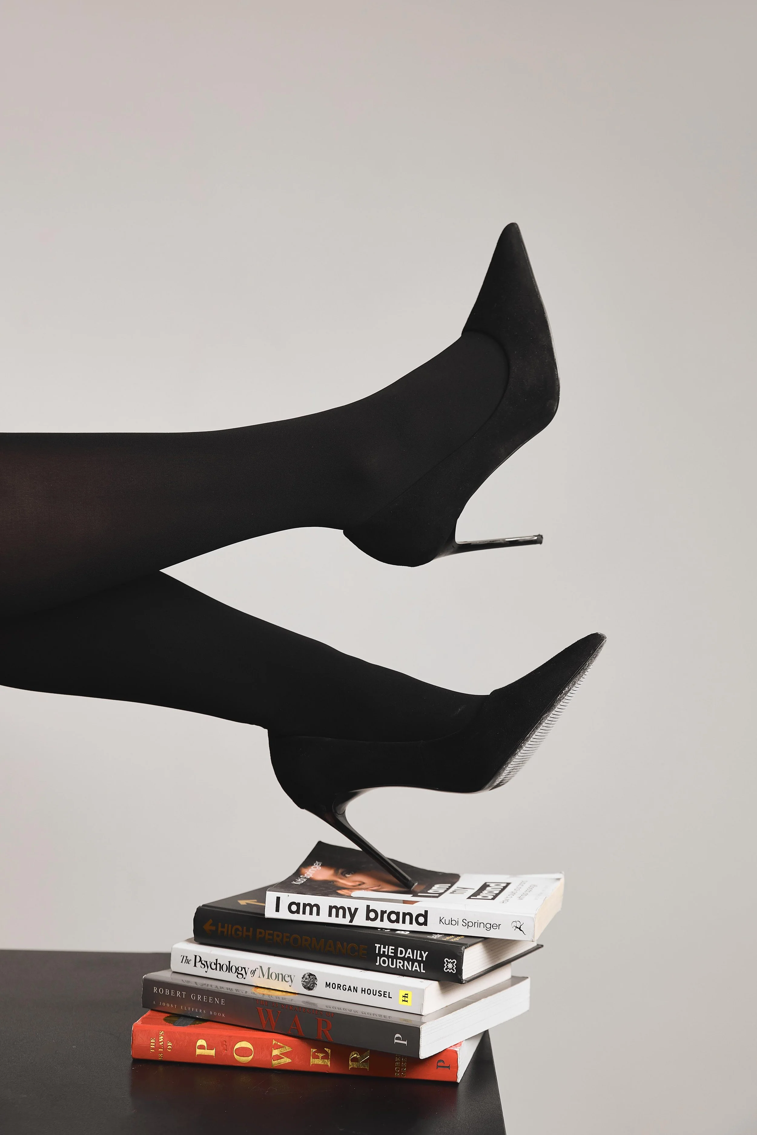 A pair of black high-heeled shoes worn with black stockings, balanced on a stack of magazines on a black table against a plain background.