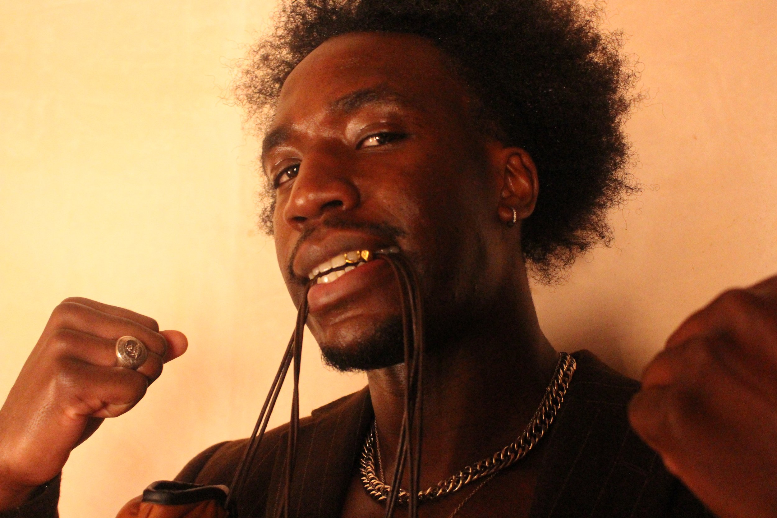 A young man with dark skin, curly hair, and tattoos on his arm is holding a pair of sunglasses near his mouth. He is wearing a silver chain necklace, small hoop earrings, and a ring on his finger, and has gold teeth visible as he smiles.