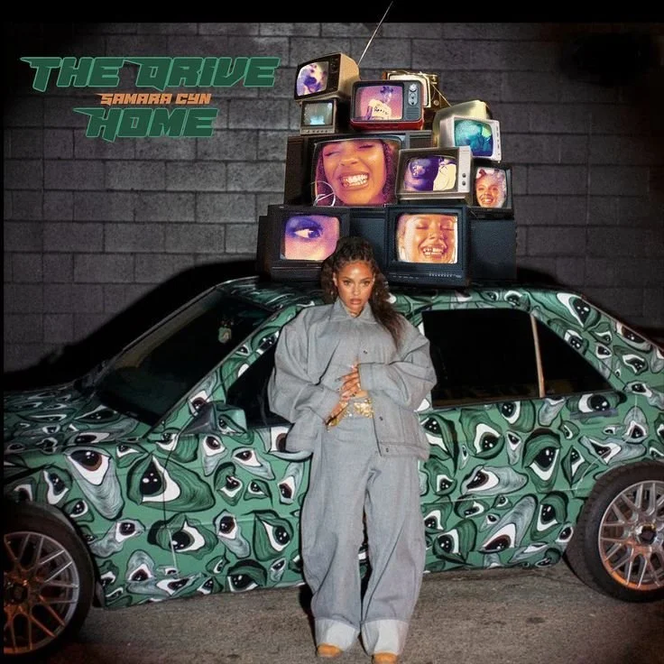 A woman in gray outfit standing in front of a camo-patterned sports car with multiple stacked vintage televisions on top displaying faces. There is a green and orange text logo saying 'The Drive Samarra Cyn Home' on a dark brick wall.