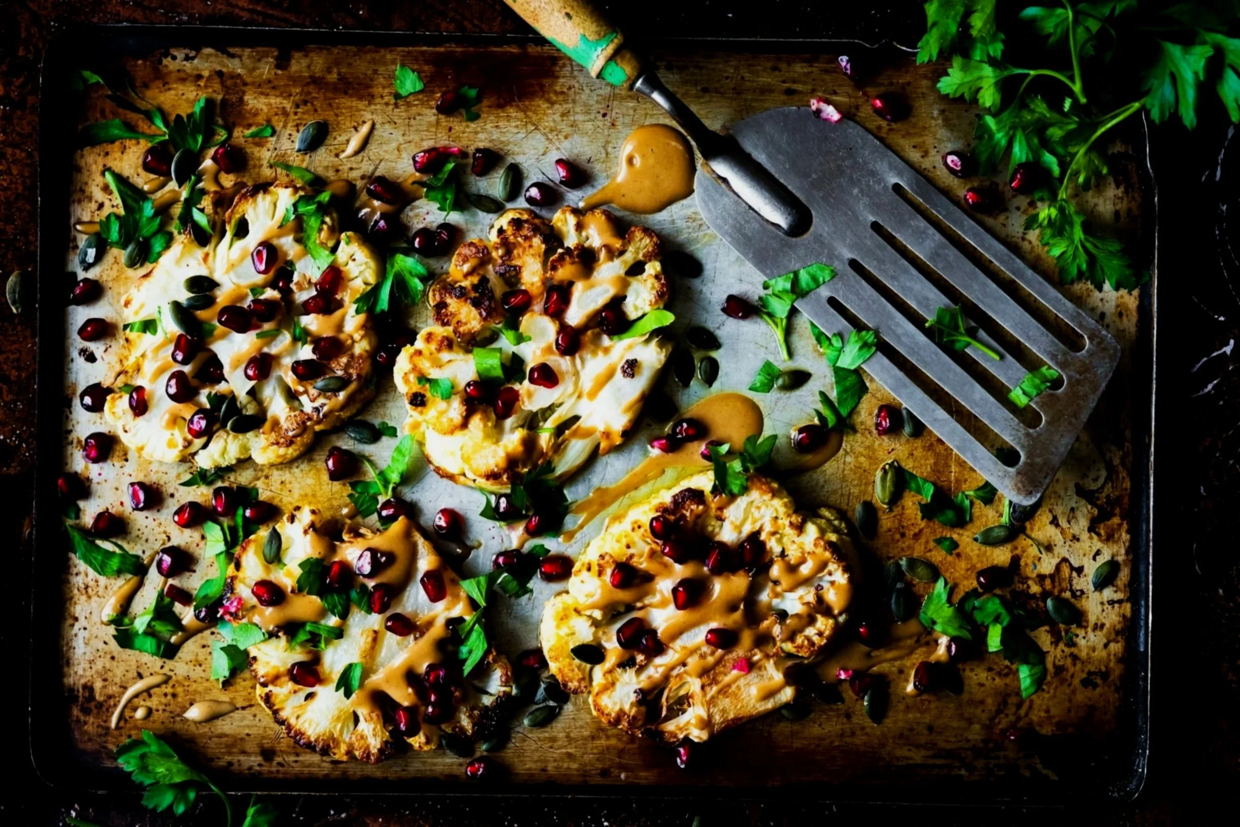 Roasted cauliflower steaks topped with pomegranate seeds, chopped herbs, and drizzled with sauce on a baking sheet.