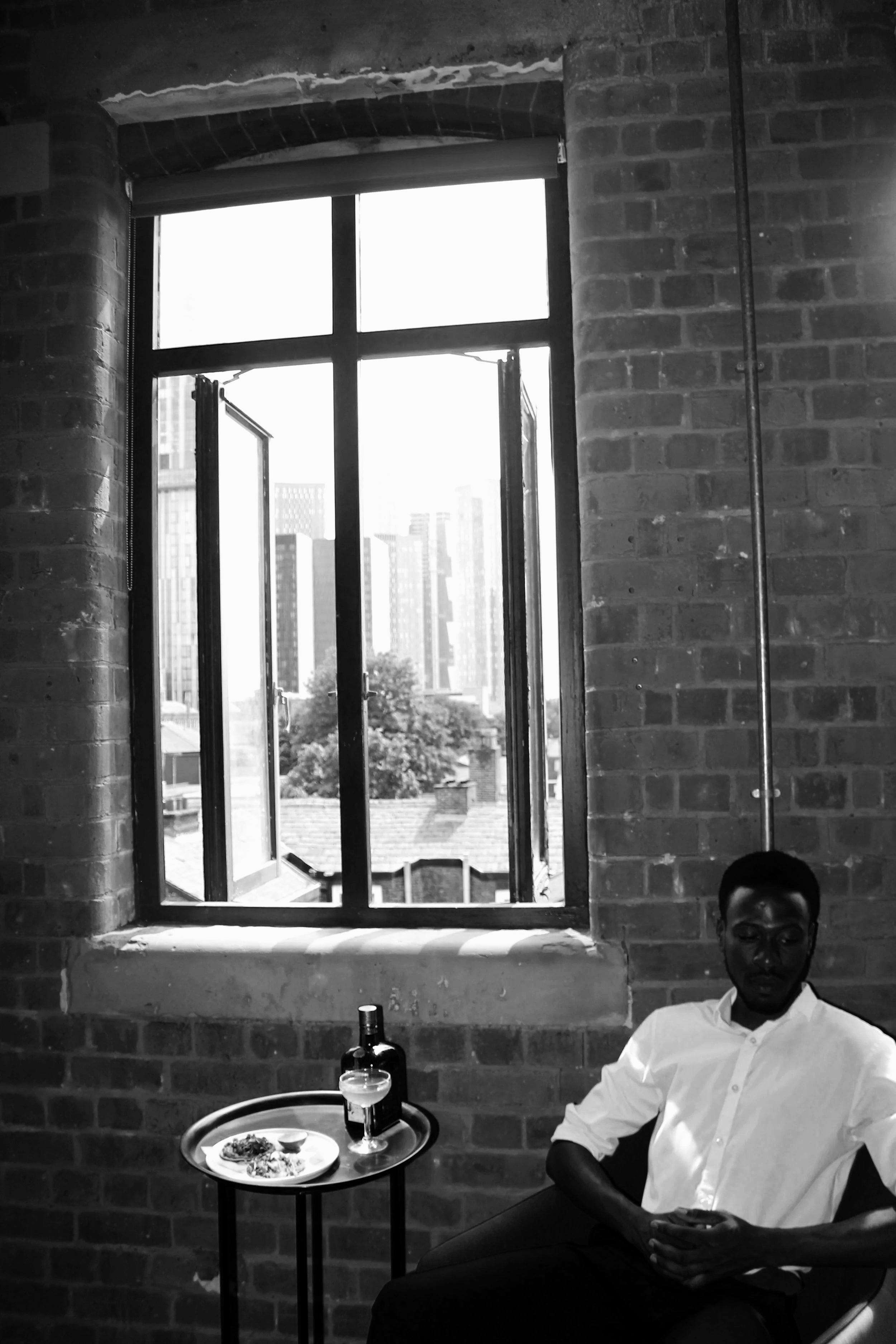A man sitting in a restaurant with an open window behind him, showing tall buildings and a cityscape in the background. The man is wearing a white shirt and appears to be holding a phone. On the table next to him, there is a tray with food, a glass of wine, and a bottle.