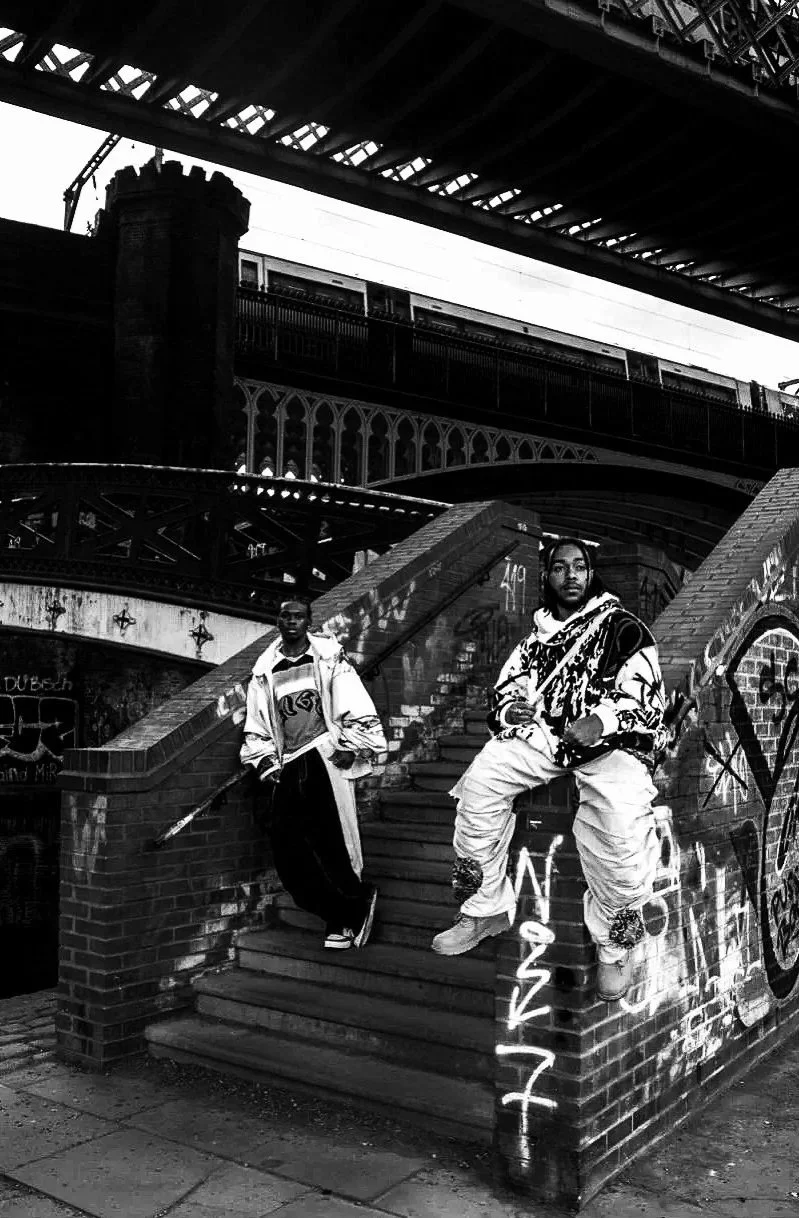 Two men stand on graffiti-covered stairs under a bridge in an urban setting, with a train passing overhead.