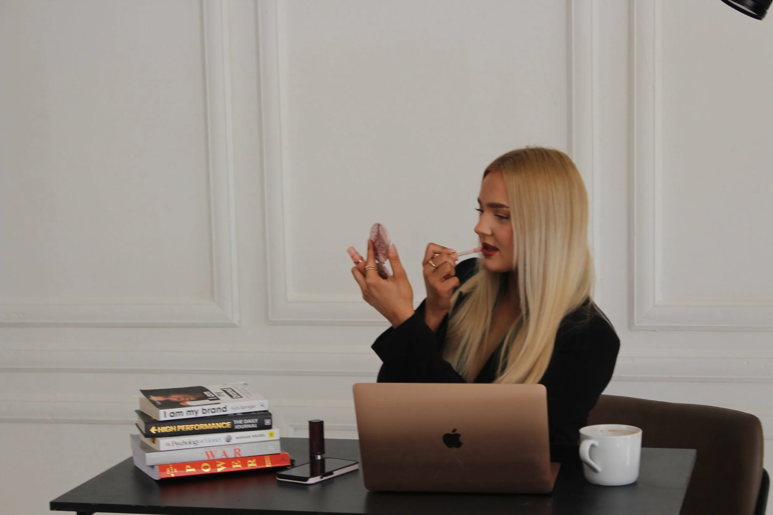 Blonde woman applying lipstick at her desk with books, a MacBook, smartphone, and coffee mug.
