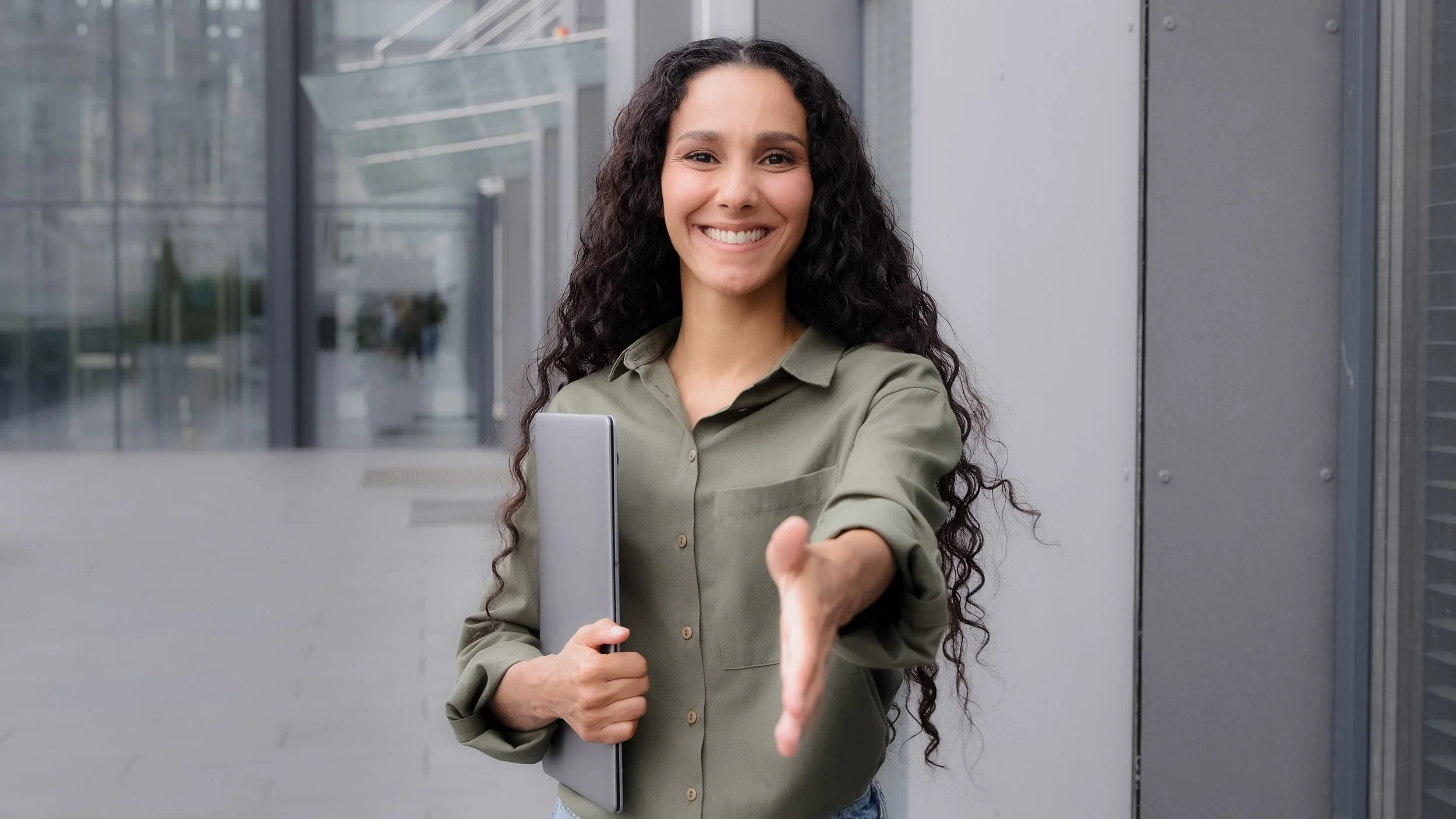 A woman with long curly hair smiling and extending her hand for a handshake outside a modern building, holding a laptop.