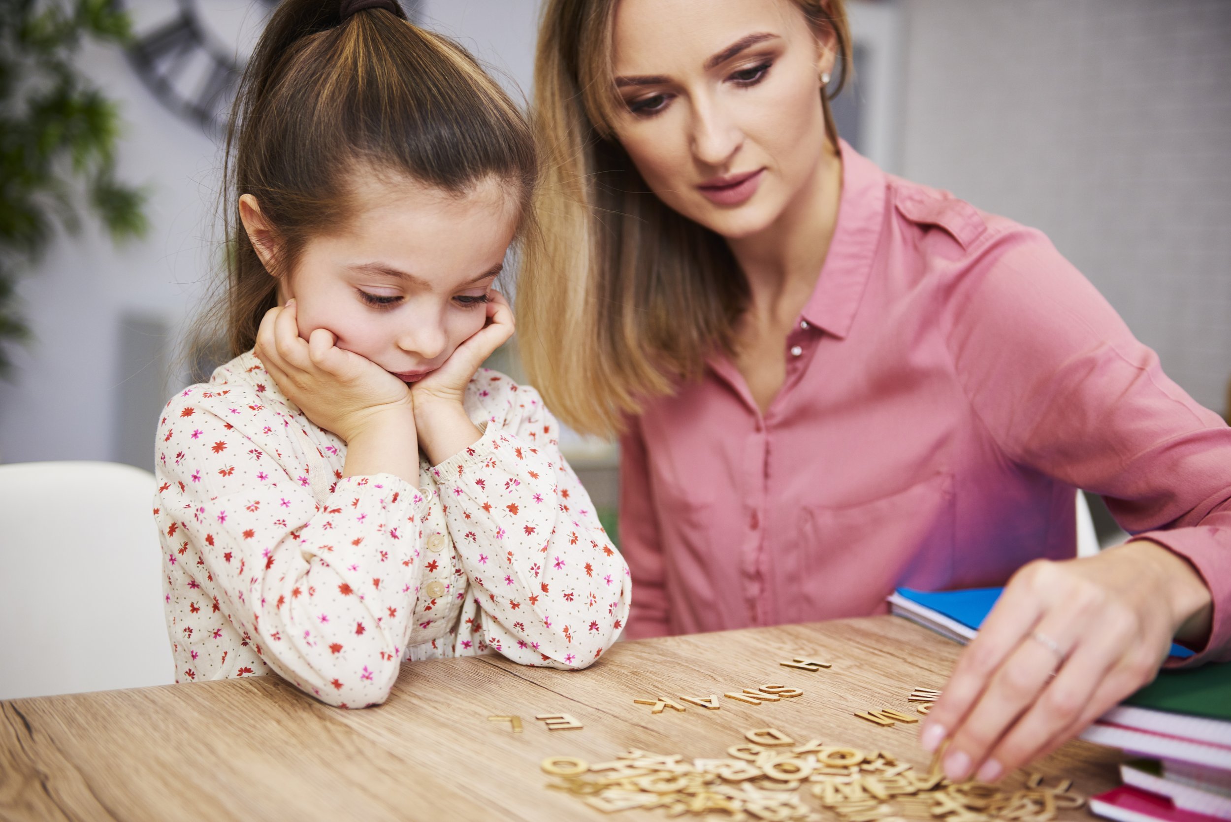 A woman and a young girl sitting at a wooden table, playing a word game with wooden letter tiles. The woman is wearing a pink blouse, and the girl is wearing a white shirt with colorful patterns. The girl is resting her chin on her hands, looking at the tiles, while the woman is reaching to pick up a tile. There are some piles of books or notebooks on the table.