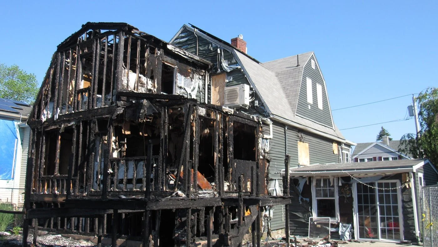 A two-story house with extensive fire damage, especially on the left side. The wooden structure is scorched and charred, with some parts blackened and broken. The house has siding on the right and a large window on the ground floor, with a glass sunroom attached to the side. The sky is clear and blue, and neighboring houses are visible in the background.