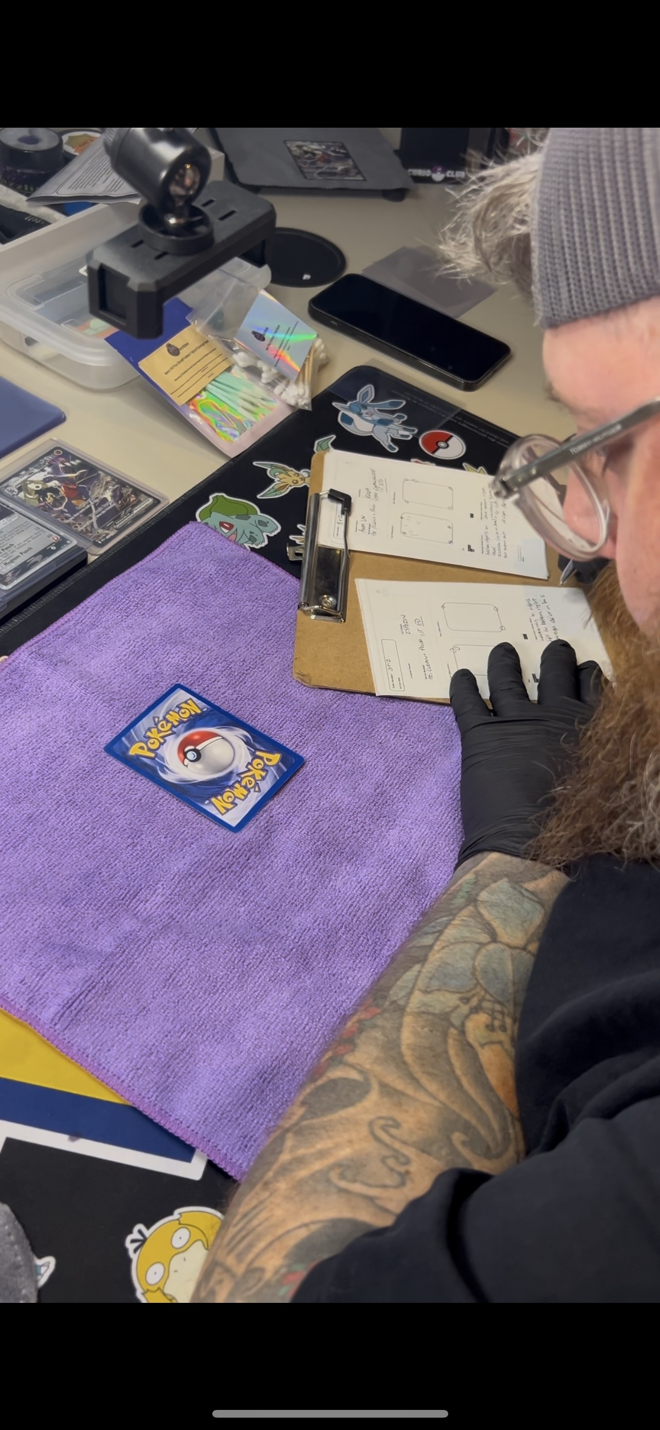 A person with tattoos on their arm and wearing black gloves is working at a cluttered table with a purple towel, Pokemon cards, stickers, a smartphone, and documents, possibly engaged in trading or collecting Pokemon cards.