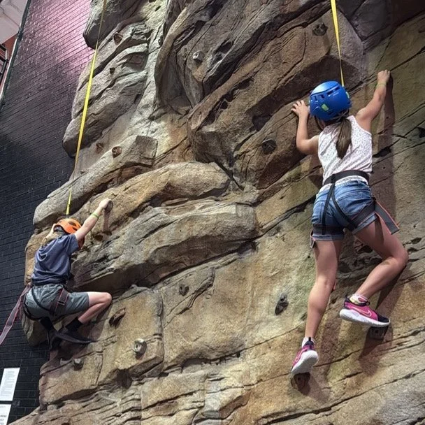 Two children rock climbing indoors on a textured wall with safety harnesses and helmets.