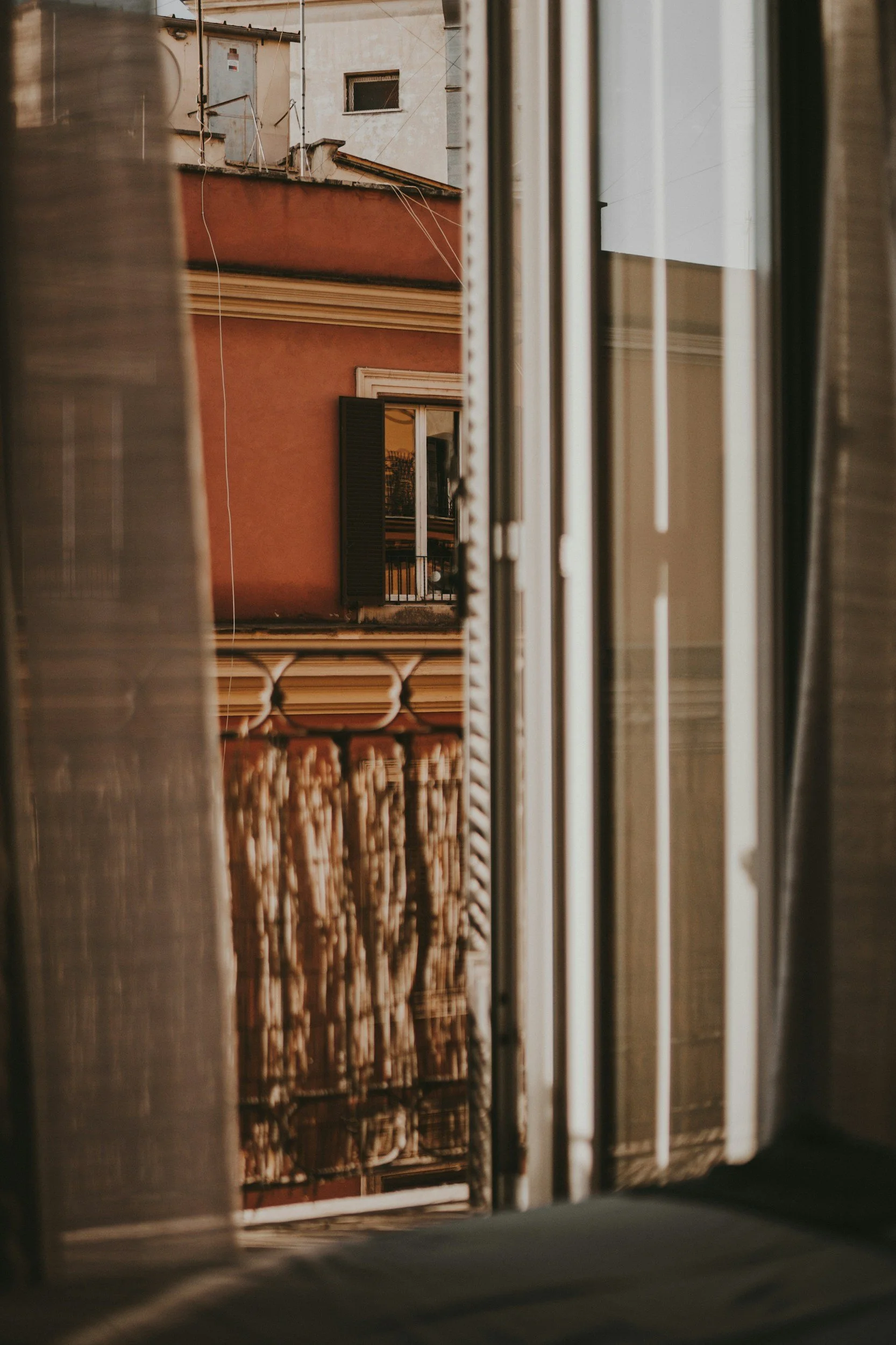 Veduta di un edificio con una finestra aperta e un balcone attraverso una finestra con tende semiaperte.