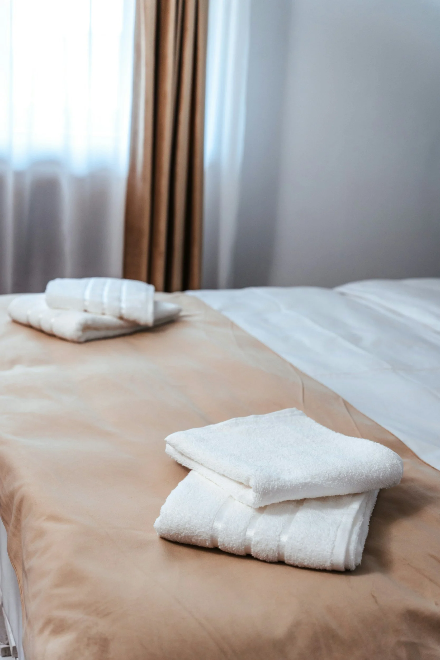 Letto con lenzuola bianche e asciugamani piegati, vicino a una finestra con tende beige.