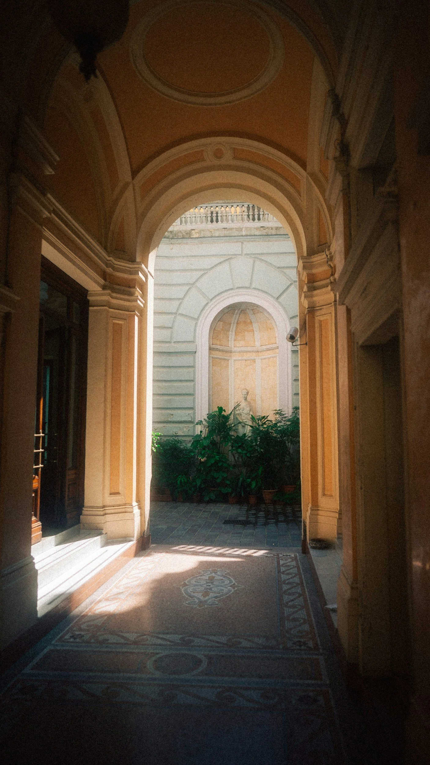 Inquadratura di un cortile interno con archi decorativi, piante verdi e una statua in fondo, illuminato dalla luce naturale.