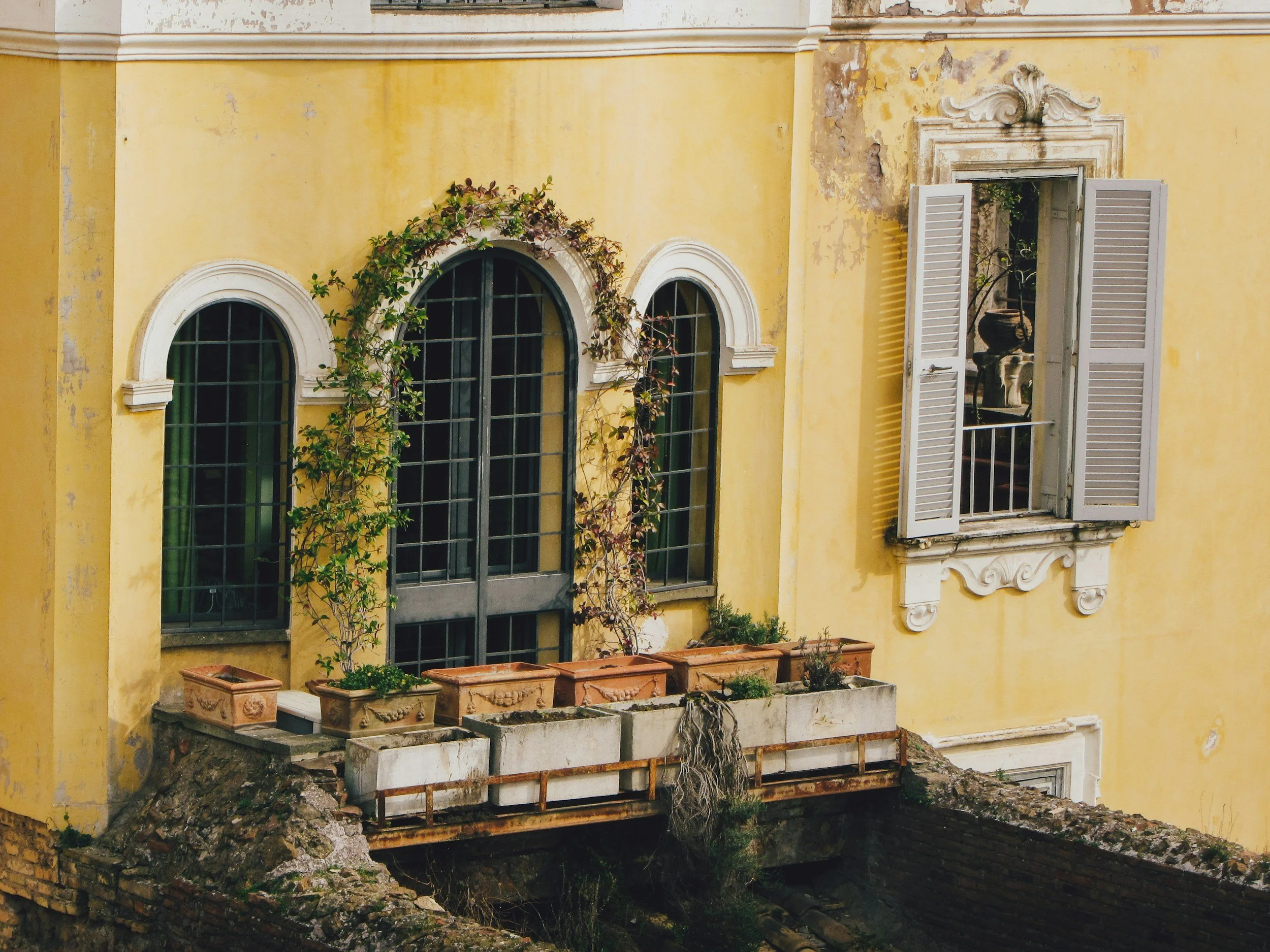 Facciata di edificio con pareti gialle, tre finestre con grate verdi e una finestra con persiane blu aperte e decorazioni in rilievo sopra.