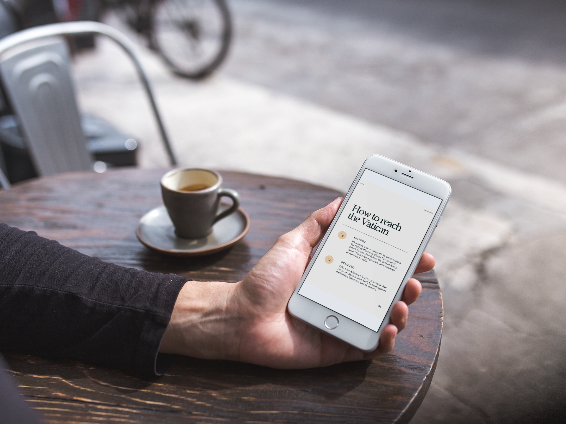 Persona seduta a un tavolo di legno all'aperto con una tazza di caffè e un telefono cellulare che mostra un articolo intitolato "How to reach the Vatican".