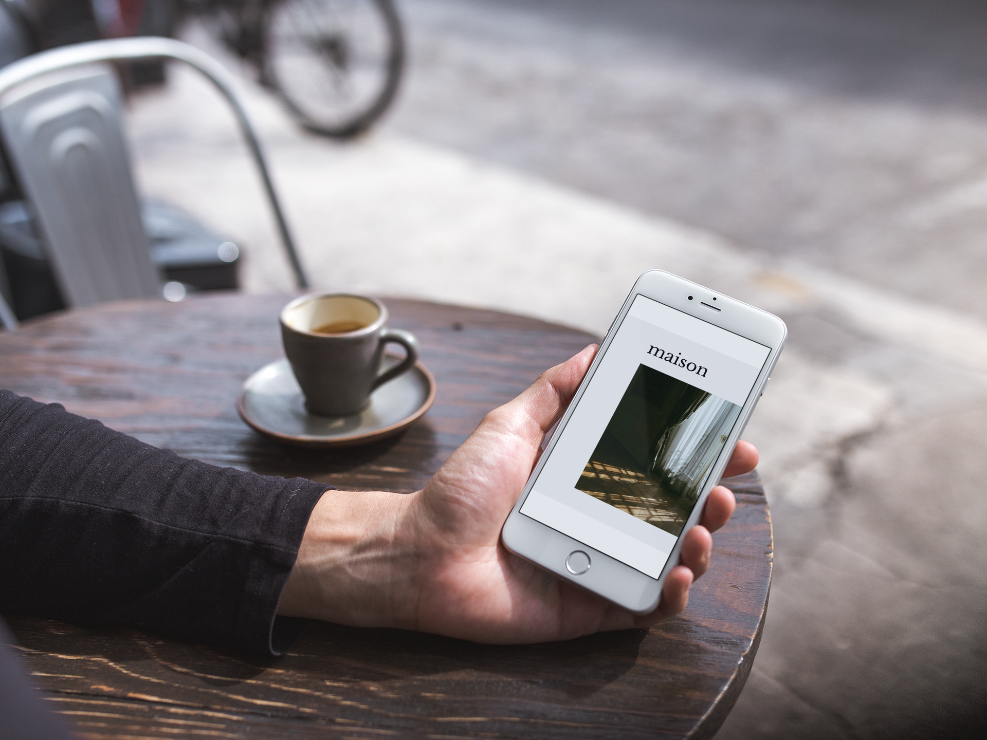 Persona che tiene uno smartphone con la schermata che mostra la parola 'maison' e una foto di un interno con tende e luce naturale, seduta a un tavolo di legno con una tazza di caffè o tè.