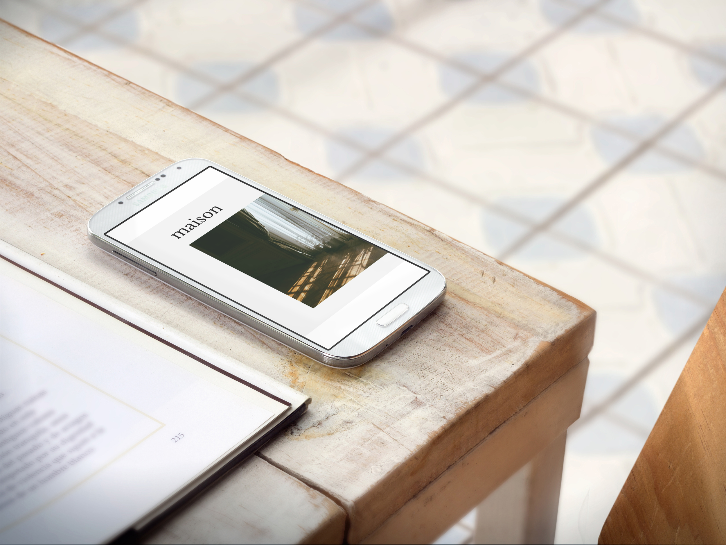 Smartphone bianco con schermo acceso che mostra il testo 'maison' e una foto di una stanza, appoggiato su un tavolo di legno con un libro aperto vicino.