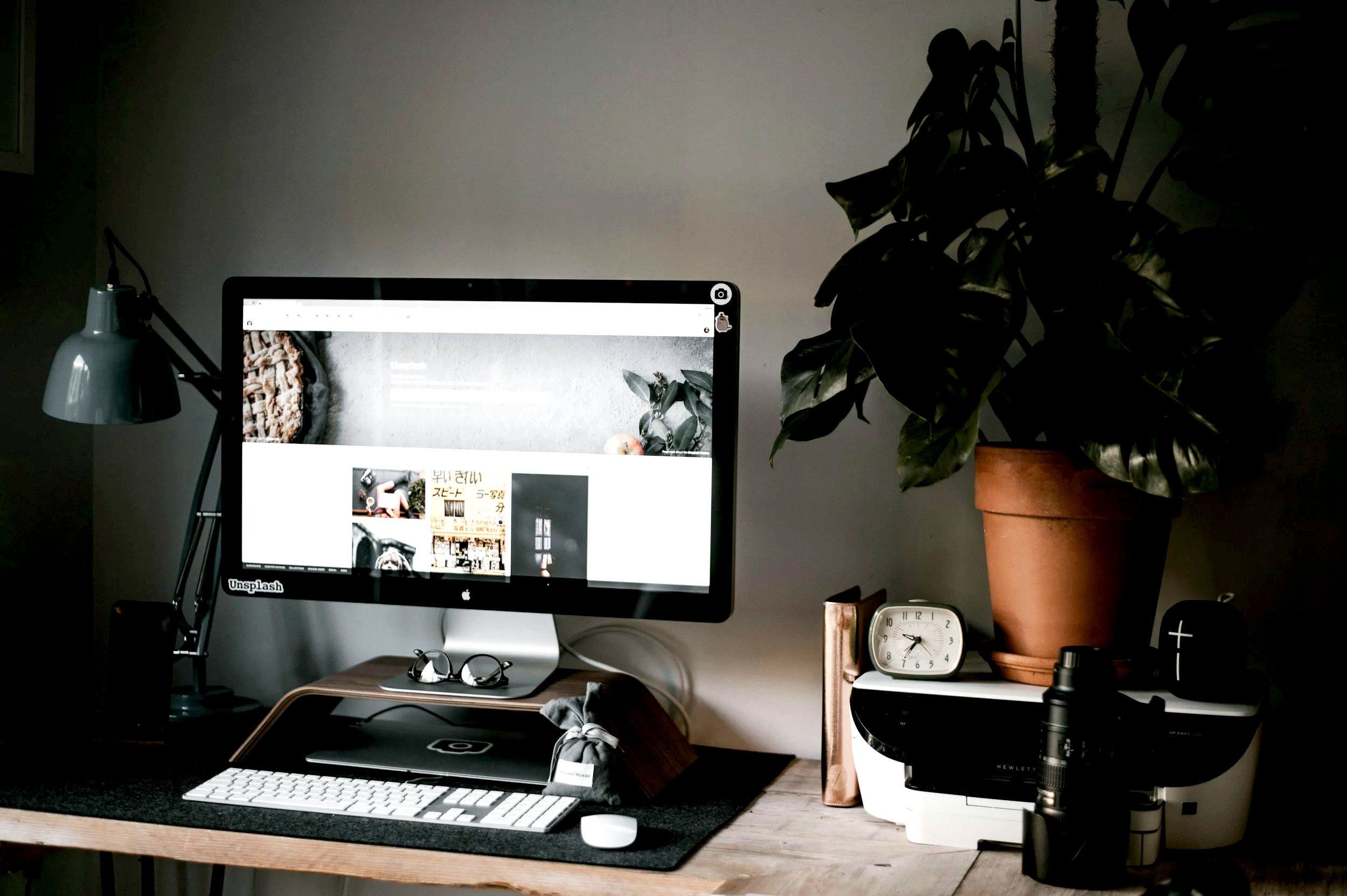 Scrivania con computer, lampada da tavolo, occhiali, taccuino, orologio e una grande pianta in vaso