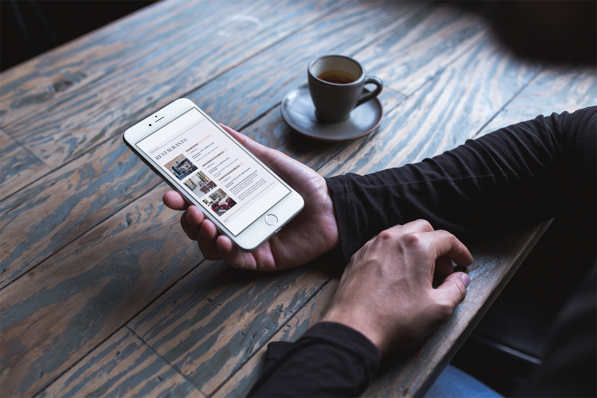 Persona che utilizza uno smartphone con una pagina web di ristoranti aperti, seduta a un tavolo di legno con una tazza di caffè sopra.