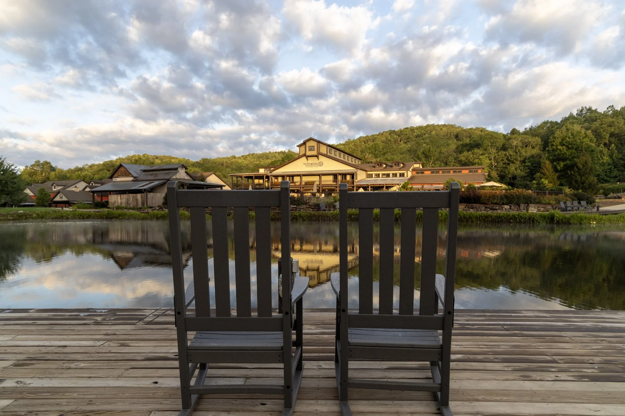 Two empty chairs on a wooden dock facing a calm lake with a large building and green hillside in the background, under a partly cloudy sky.