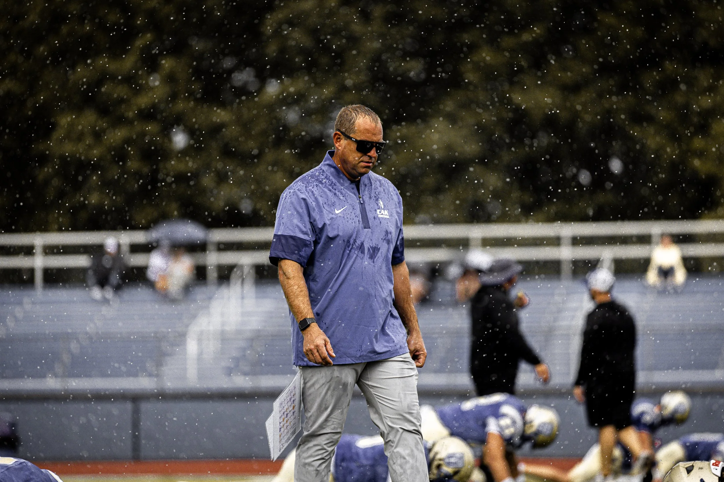 A football coach walking on the sidelines during a game in rainy weather, wearing sunglasses, a blue jacket, and gray pants, with players kneeling and standing on the field in the background.
