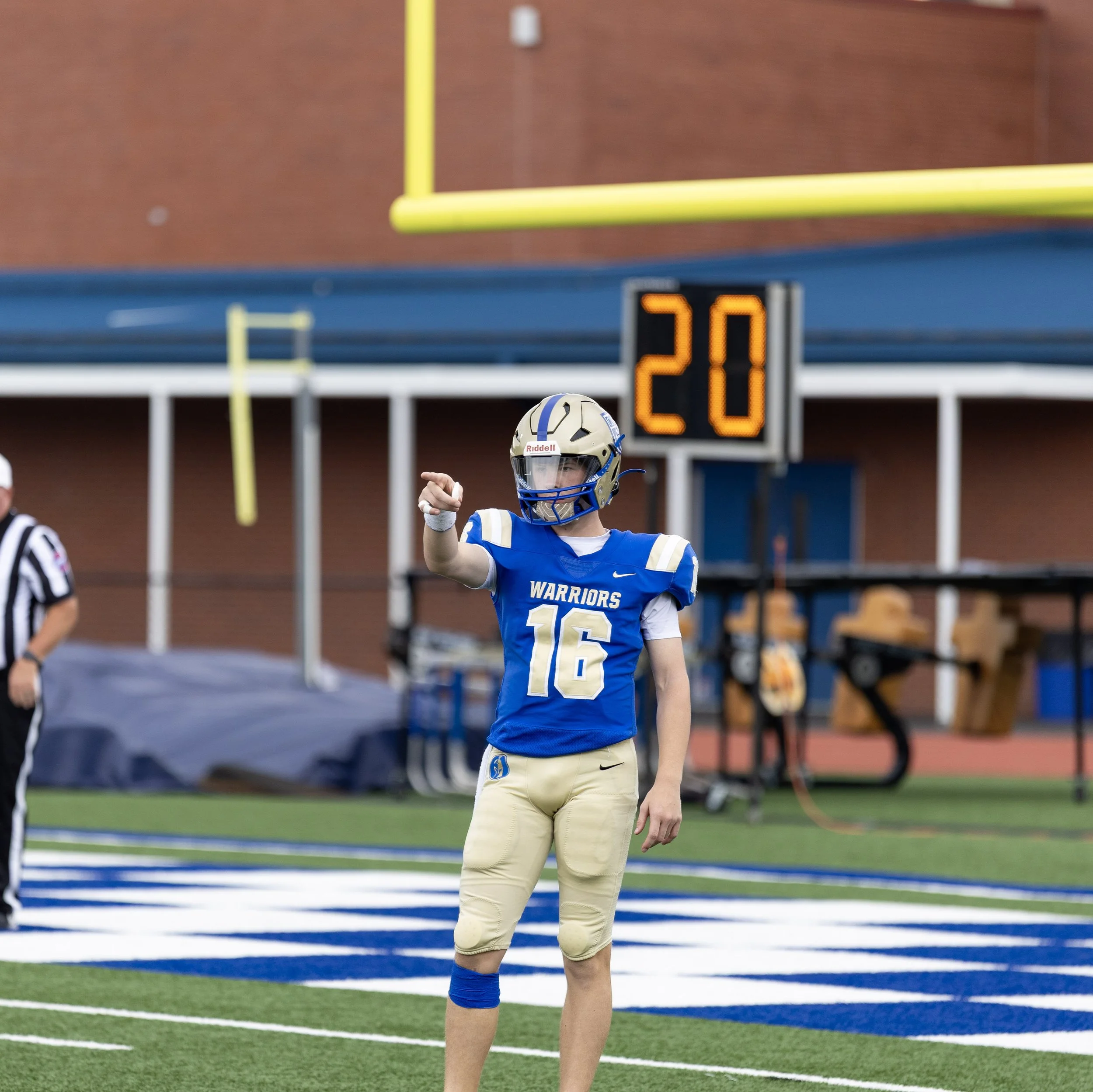 A football player wearing a blue and gold uniform with the number 16 and the team name 'Warriors' is standing on a football field. The player is pointing with their right hand and wearing a helmet. There is a digital scoreboard showing the number 20 in the background.