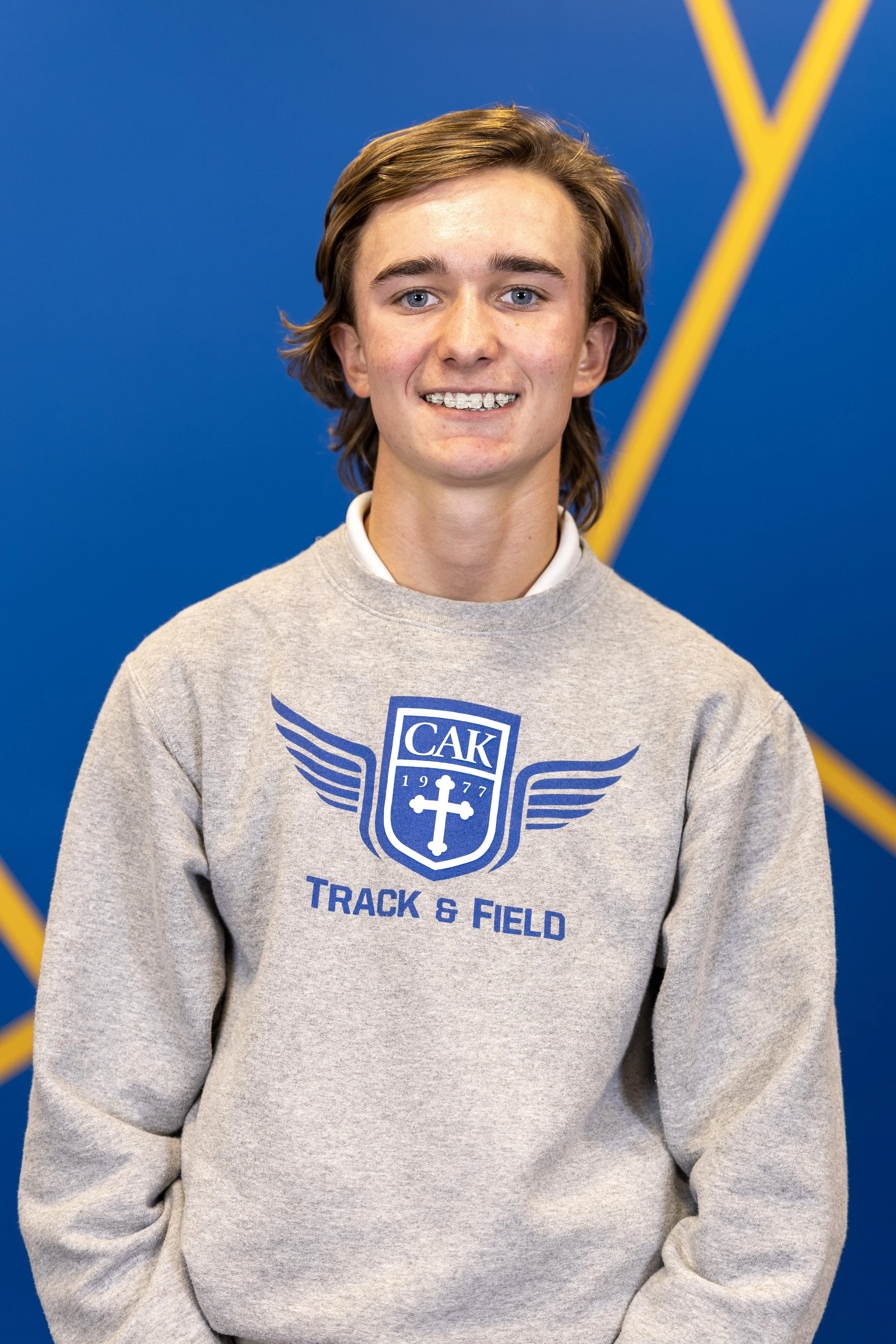 A young man with shoulder-length brown hair, wearing a gray sweatshirt with blue insignia that reads "CAK Track & Field," standing in front of a blue background with yellow lines.