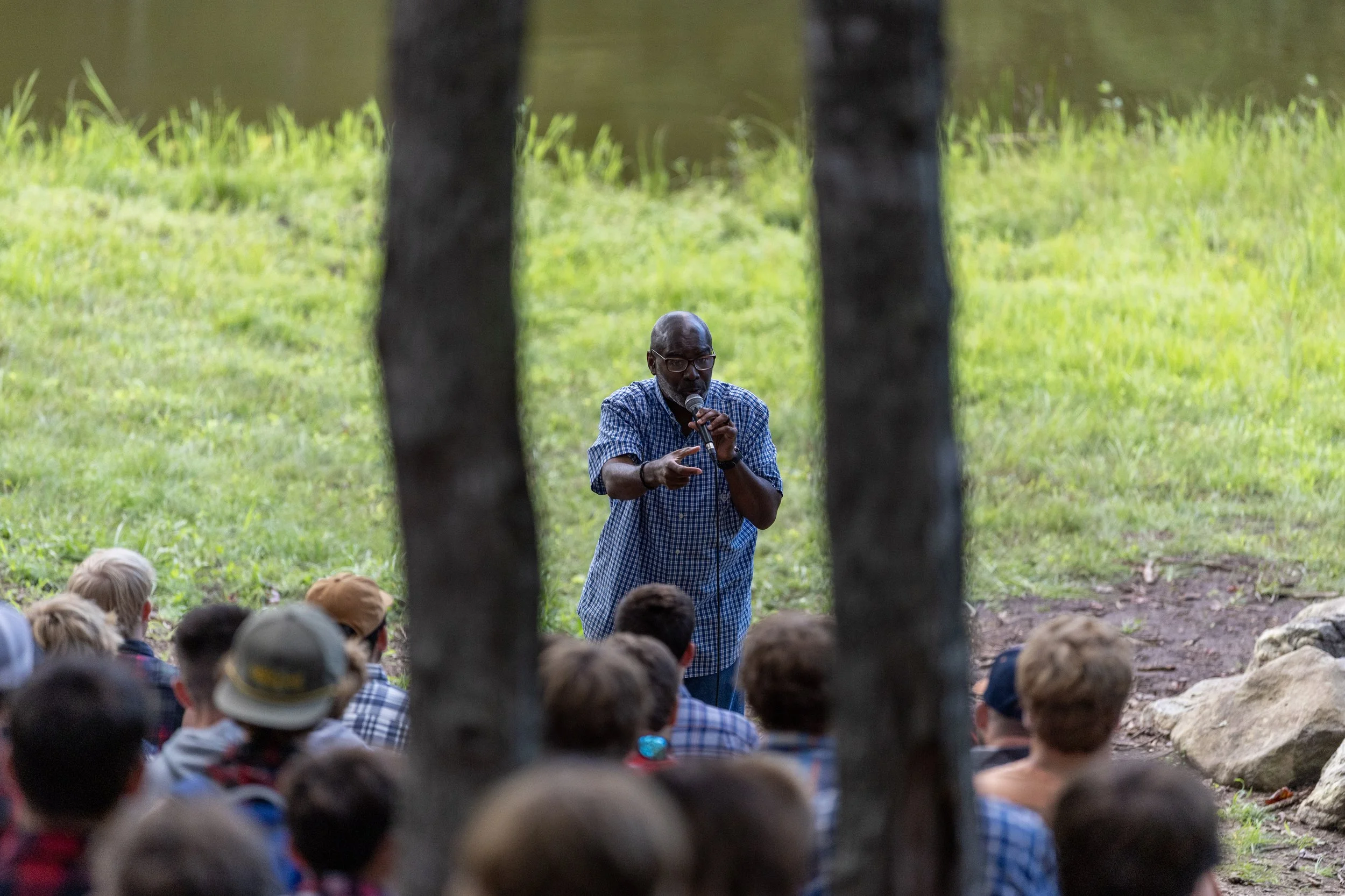 A man speaking into a microphone to a group of children seated outdoors on grass, framed by two tree trunks.