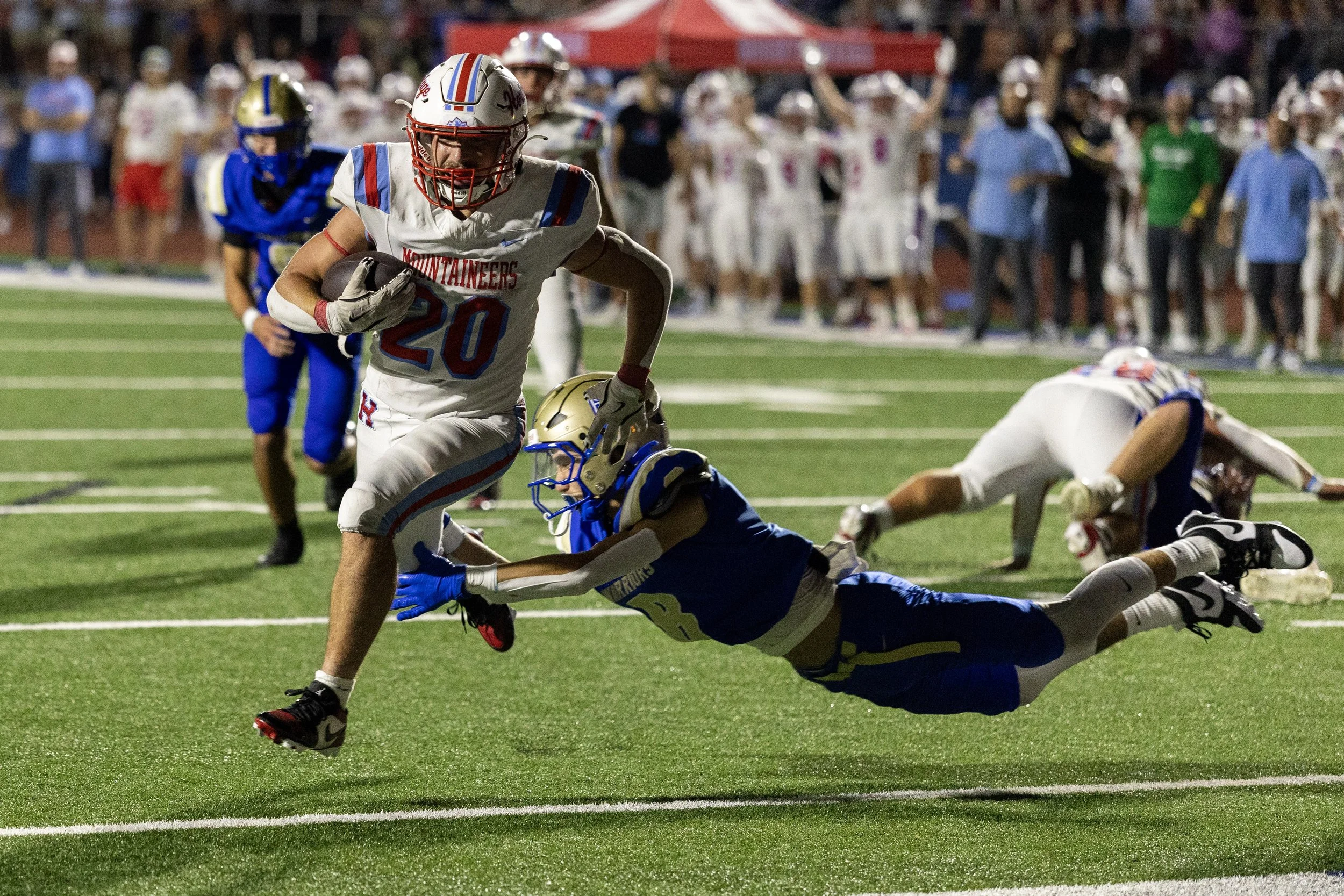 A football player in a white uniform with red and blue accents running with the ball while a player in a blue uniform dives to tackle him during a game on a lit football field at night.