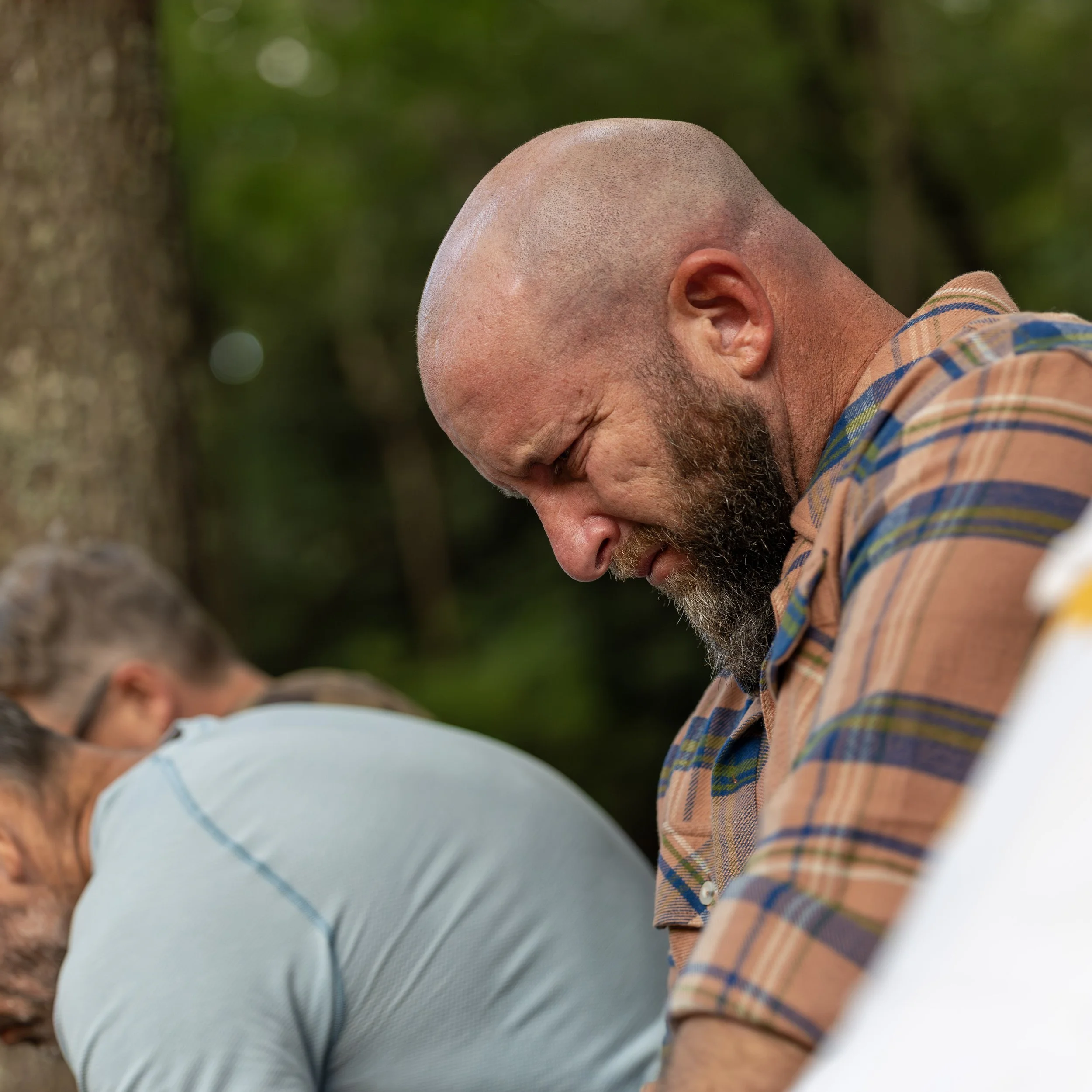 Man with a beard and a plaid shirt, eyes closed, in a forest setting, appears to be praying or meditating.