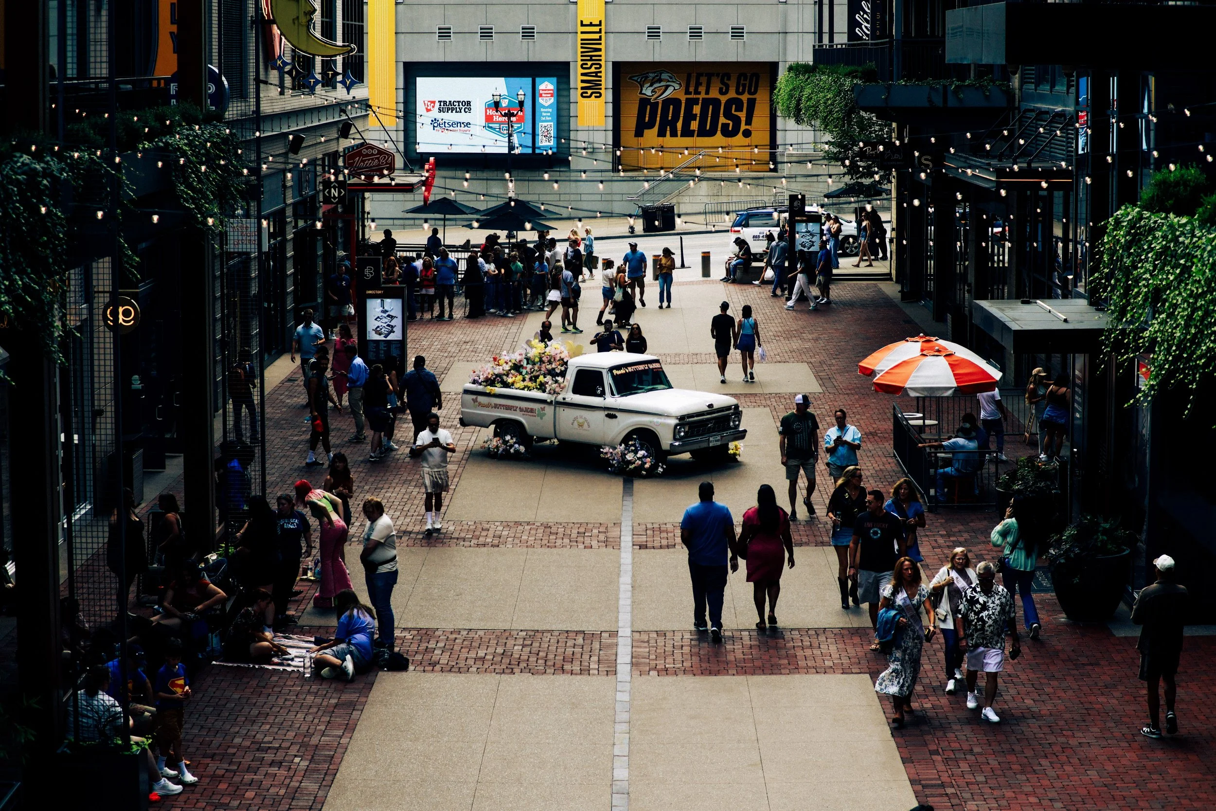 A busy outdoor area with people walking and sitting, a vintage white pickup truck decorated with flowers in the center, and various signs and umbrellas. There are businesses and advertisements visible in the background.