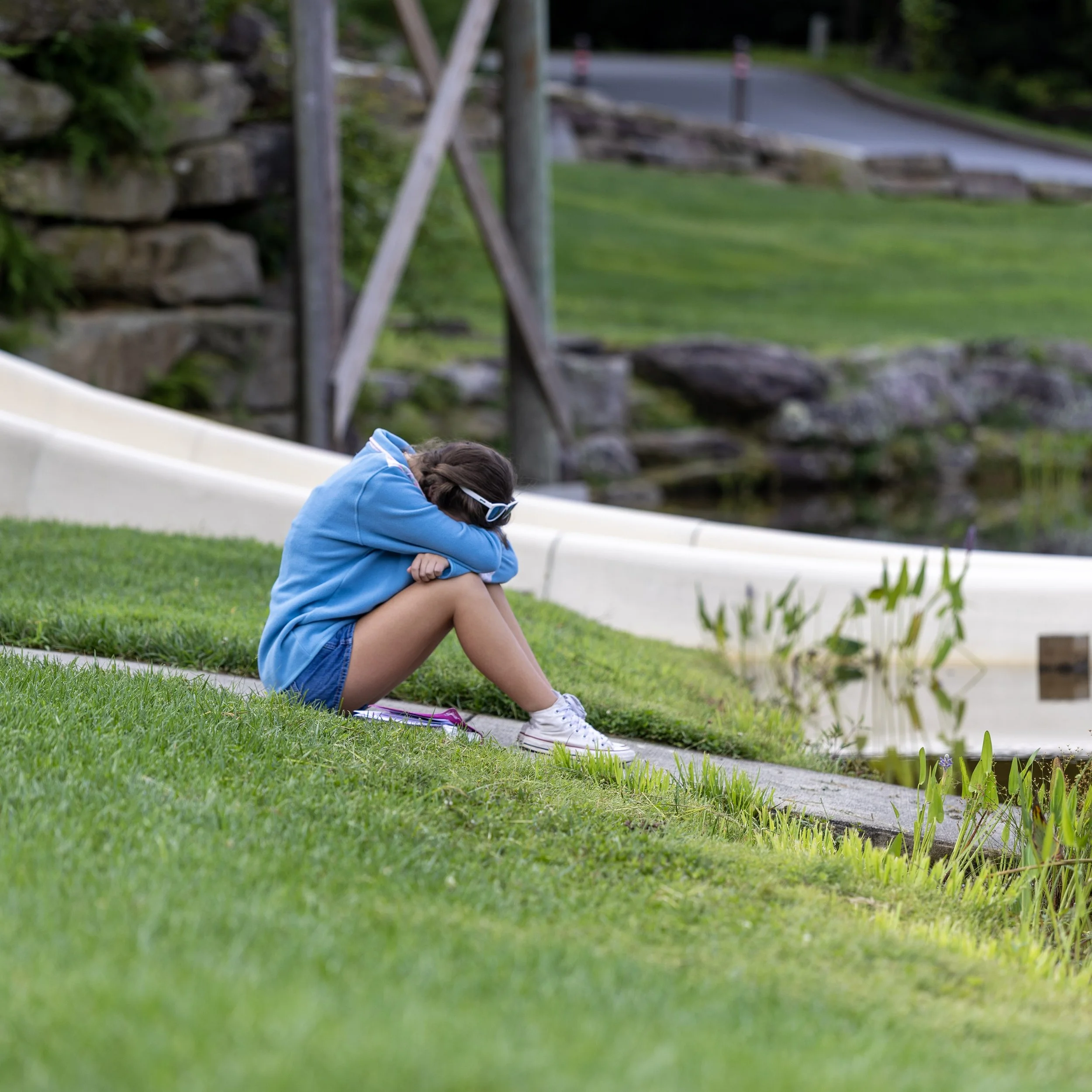 A girl sitting on the grass near a pond, hugging her knees with her head resting on her arms, wearing a blue hoodie, shorts, sunglasses, and white sneakers.