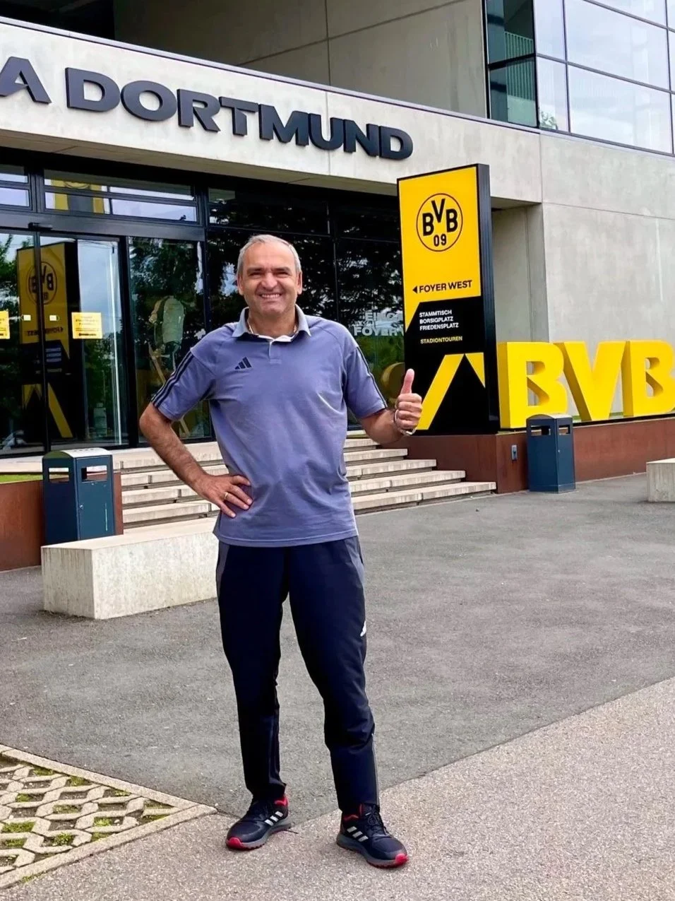 A man in athletic clothing standing in front of Borussia Dortmund stadium, smiling, with a thumbs up gesture.