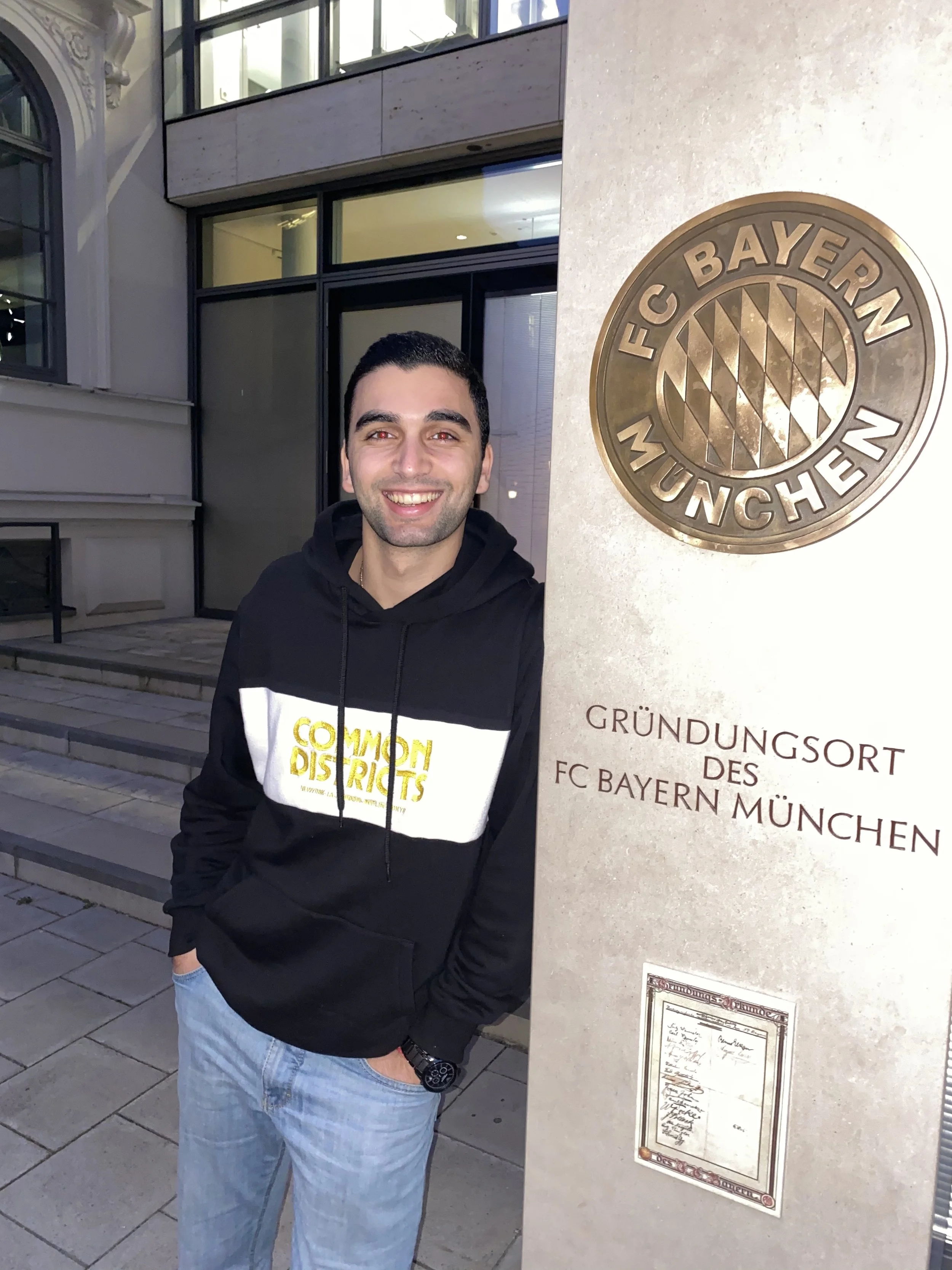 A young man standing next to a stone sign with a FC Bayern München logo and German text, smiling at the camera outside a building with large windows.