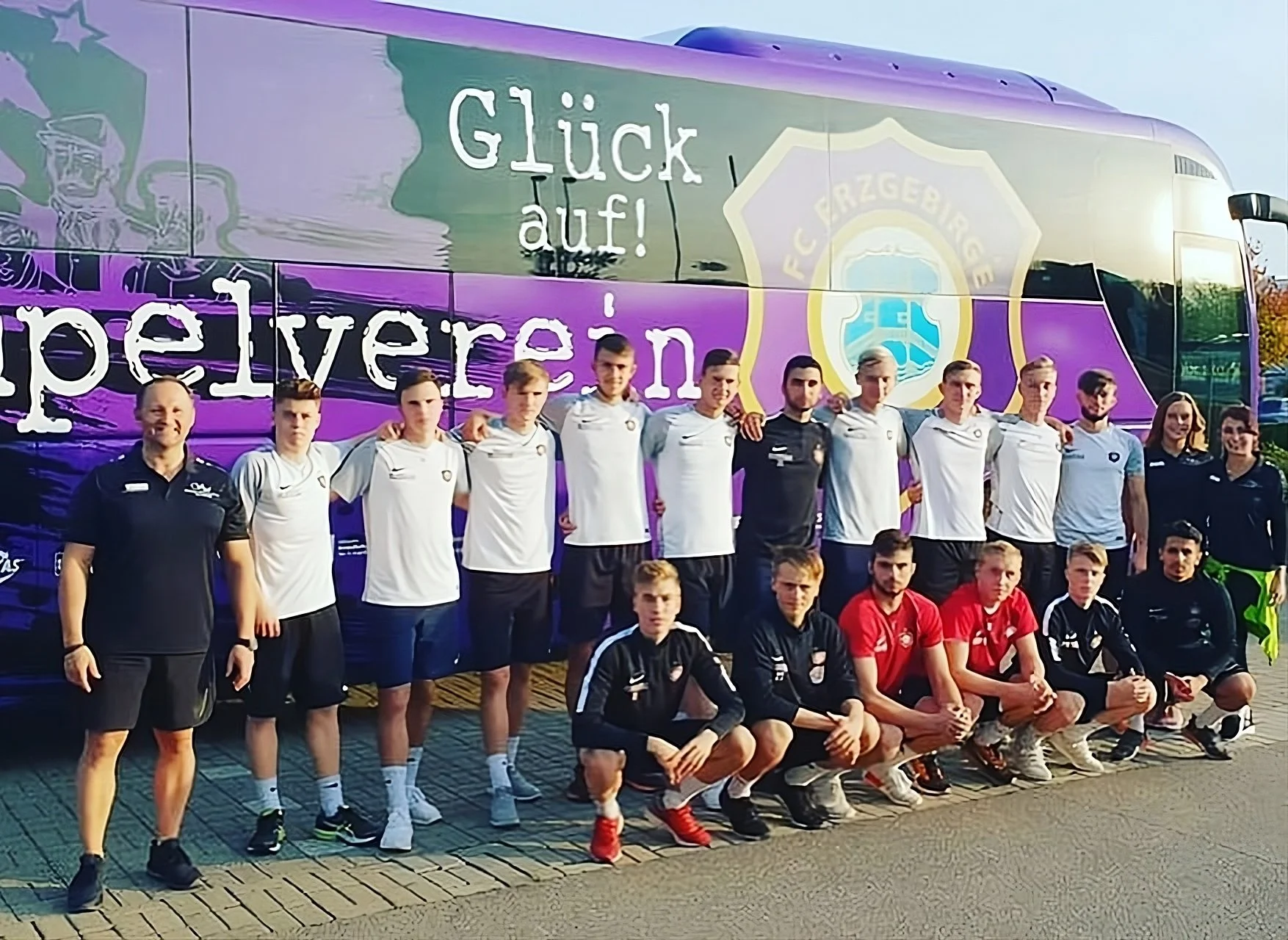A group of young soccer players and coaches posing in front of a purple bus with the FC Erzgebirge logo at sunset.