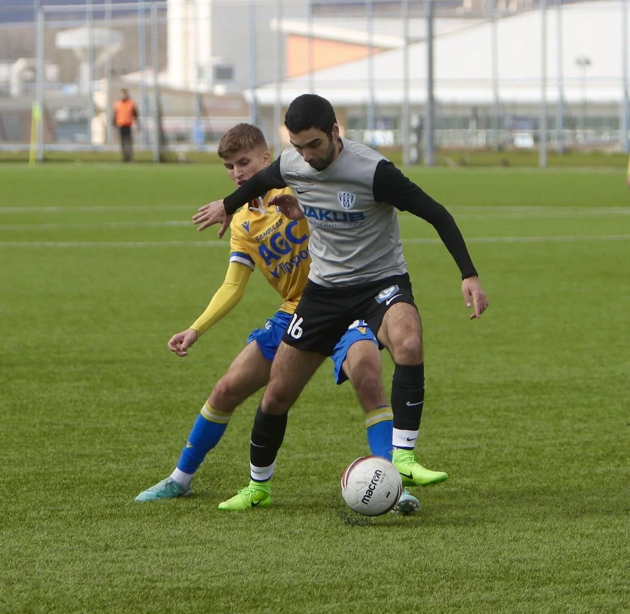 Two soccer players compete for the ball on a field, one in a gray and black uniform and the other in a yellow and blue uniform, with a goalpost and a building in the background.