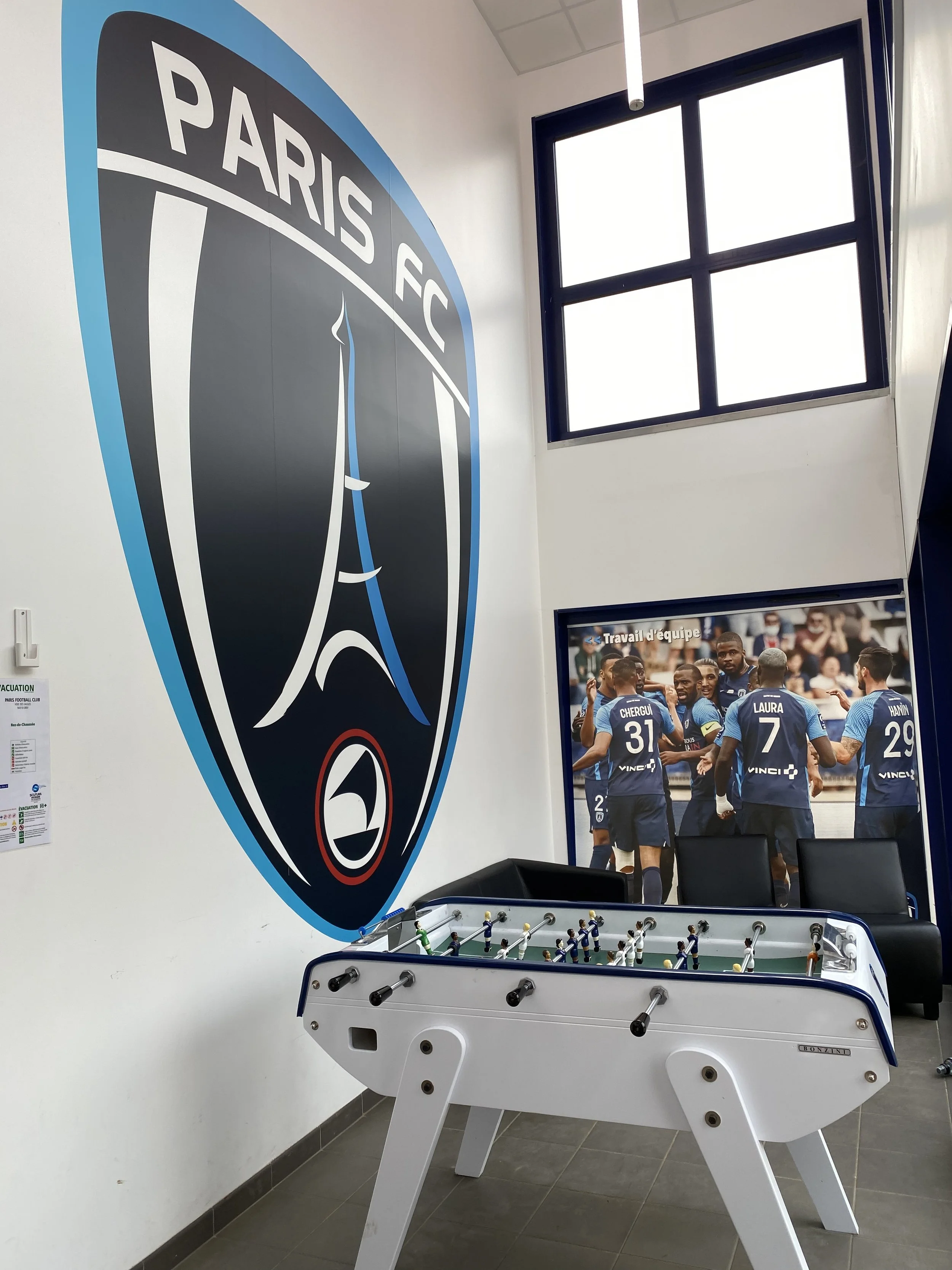 A room decorated with Paris FC football club themes, featuring a large Paris FC logo on the wall, a foosball table, and a large poster of soccer players celebrating, with some sitting on chairs by the window.