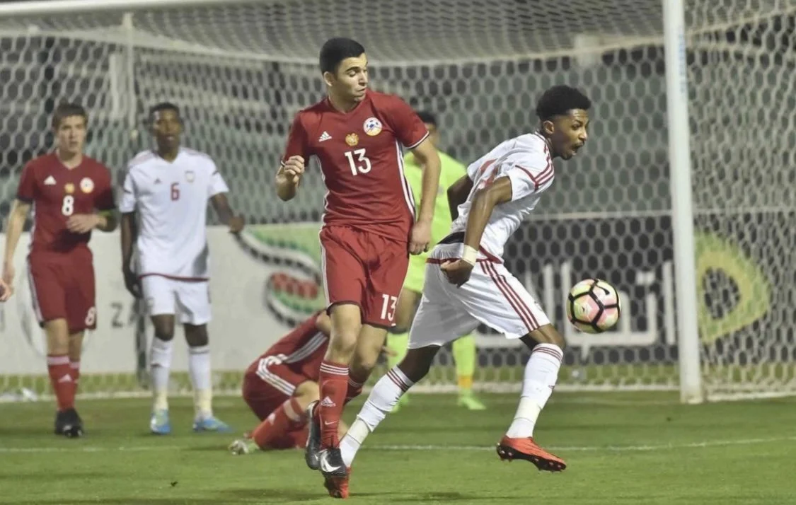 Soccer players in a game, one in a white uniform controlling the ball near the goal, others in red uniforms nearby, with a net in the background.