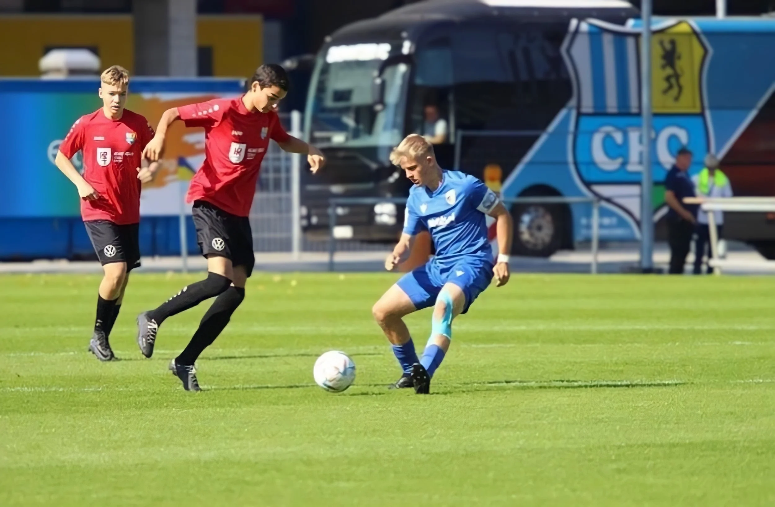Three young male soccer players on a grass field, two in red jerseys and one in a blue jersey, competing for the soccer ball during a match.