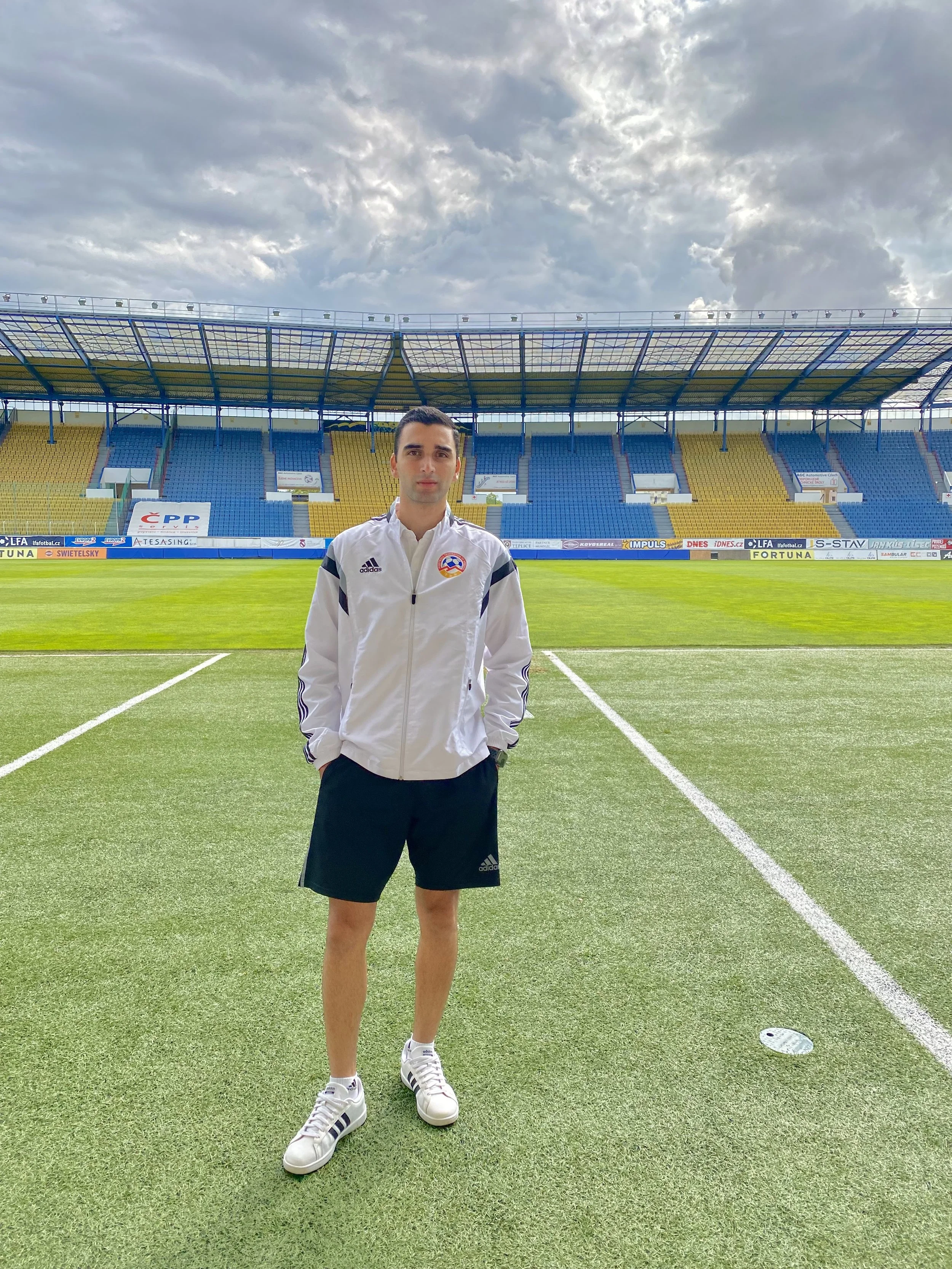 A young man in a white jacket and black shorts standing on a soccer field with a stadium in the background.