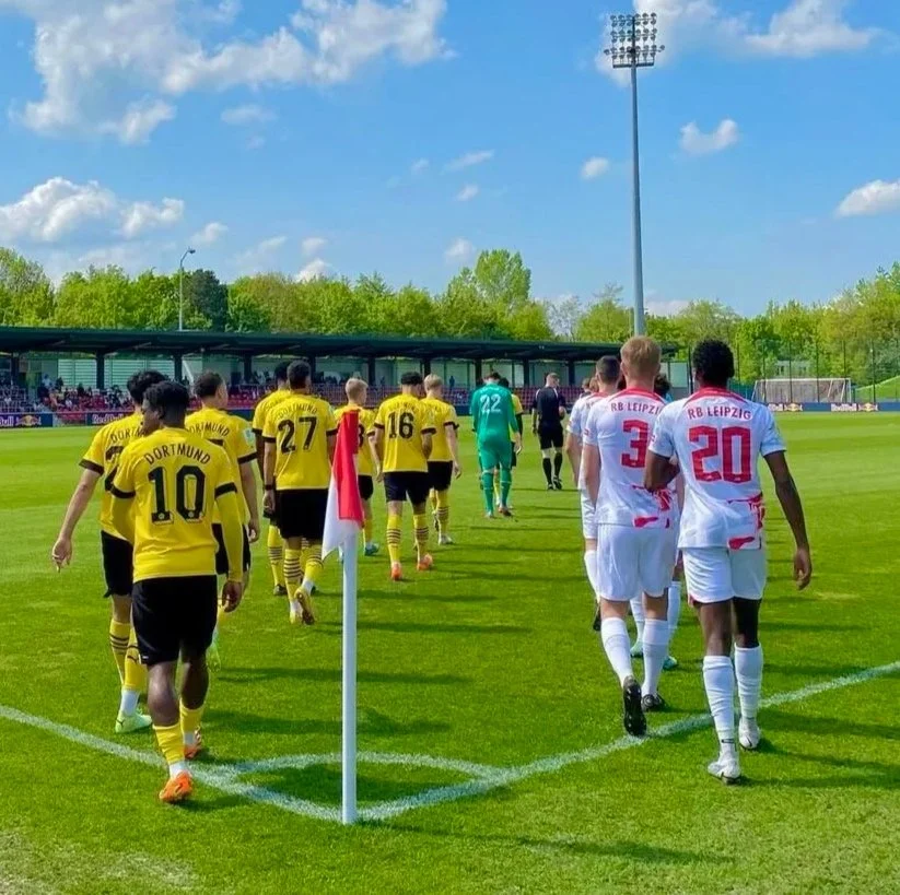 Soccer players from Borussia Dortmund and RB Leipzig walking onto the field from the corner, with a corner flag in the foreground, green grass, blue sky, and spectators in the stands.