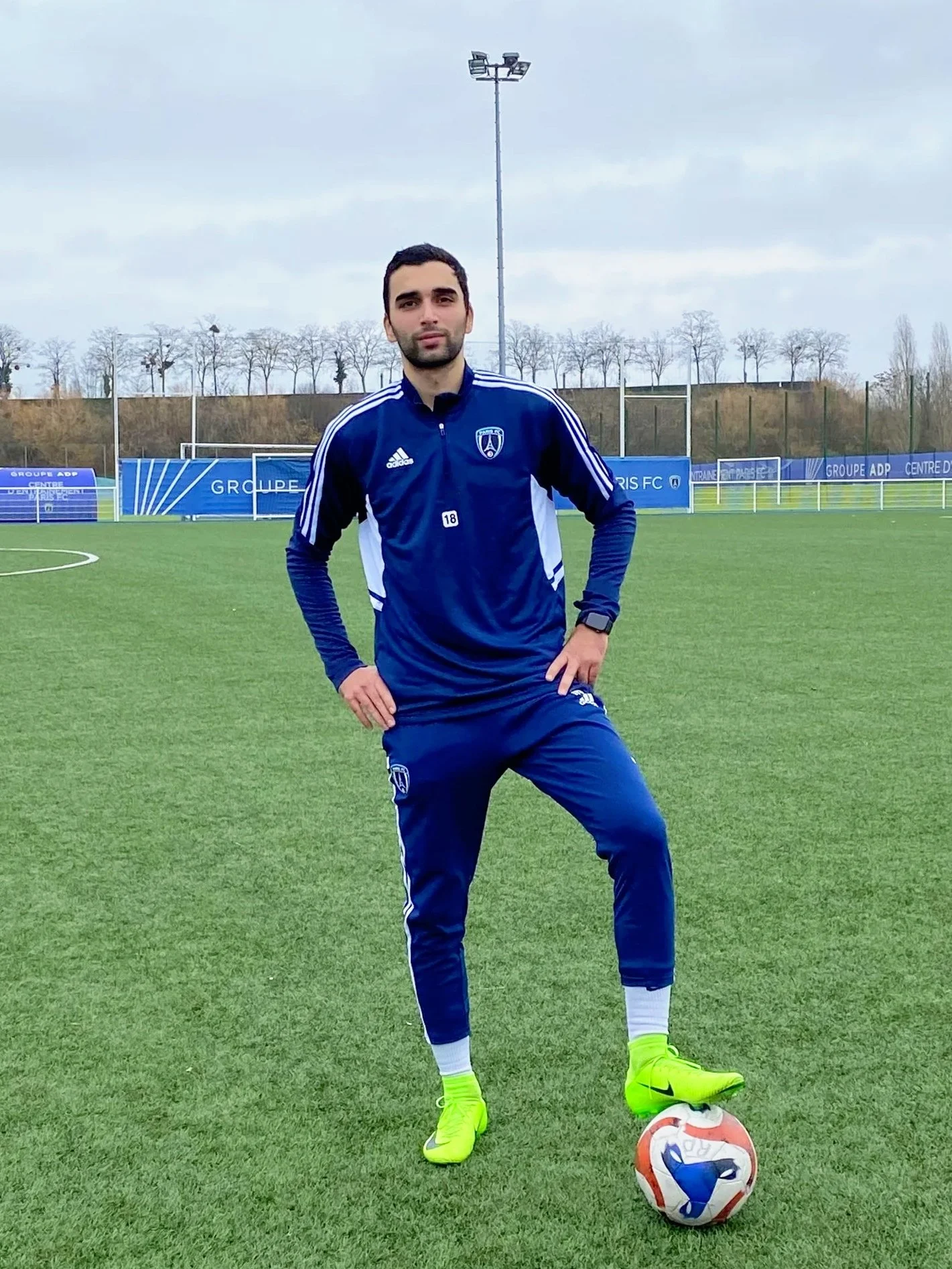 A man in a blue soccer tracksuit with white accents stands on a soccer field, with one foot on a colorful soccer ball, facing the camera with a neutral expression. The field is outdoors with goalposts and advertising banners in the background.